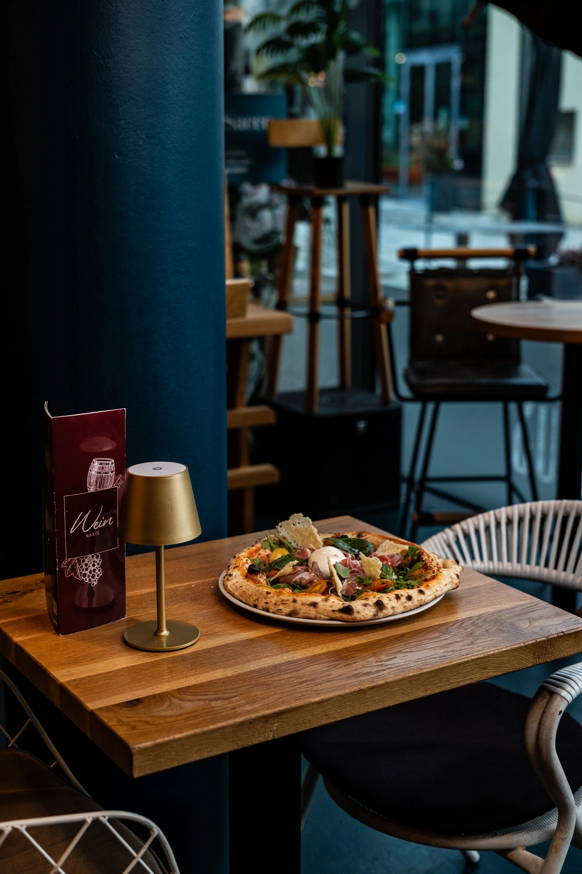 Pizza auf einem Holztablett in einem Restaurant, neben einem kleinen goldenen Lampen und einer Menükarte, im Hintergrund sind einige Stühle und Pflanzen sichtbar.