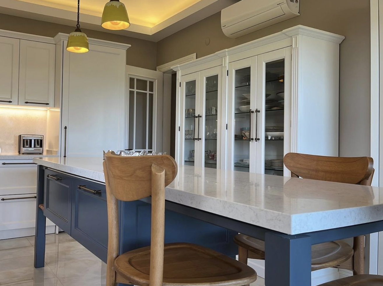 Detail view from the blue central island and dining table of an open-concept modern eclectic kitchen with rustic details, showing wall cabinets, with white cabinetry, contemporary lighting, and walnut chairs.