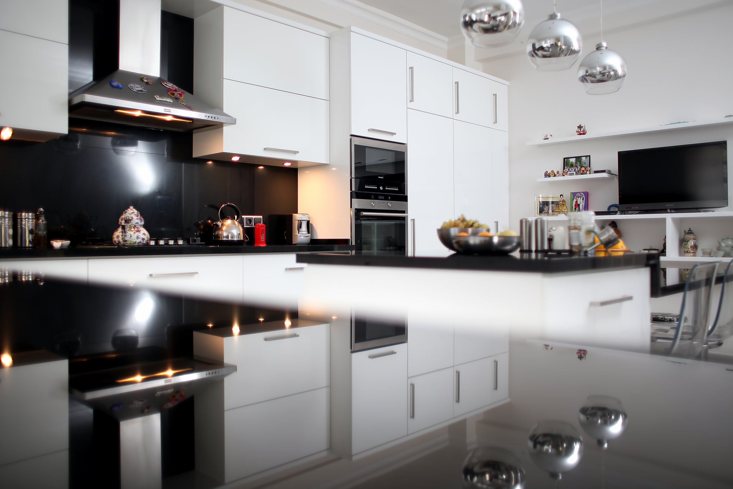 View of wall cabinets of a luxury modern kitchen with large central island, black quartz countertops, white cabinetry, contemporary lighting and high-end stainless steel appliances.