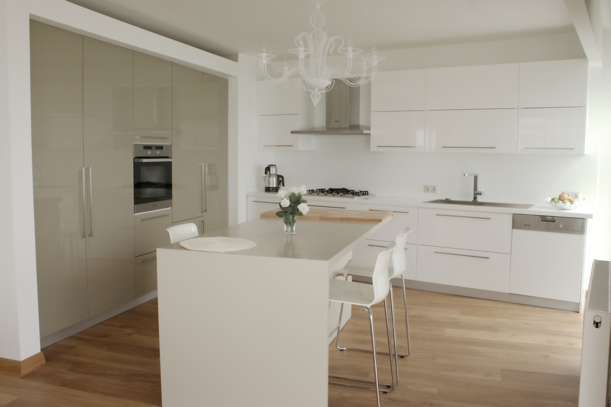 A modern minimalist kitchen with a central island and table, with grey quartz countertops, grey and white wooden cabinetry, oak hardwood floors, contemporary pendant lighting, and stainless steel appliances.
