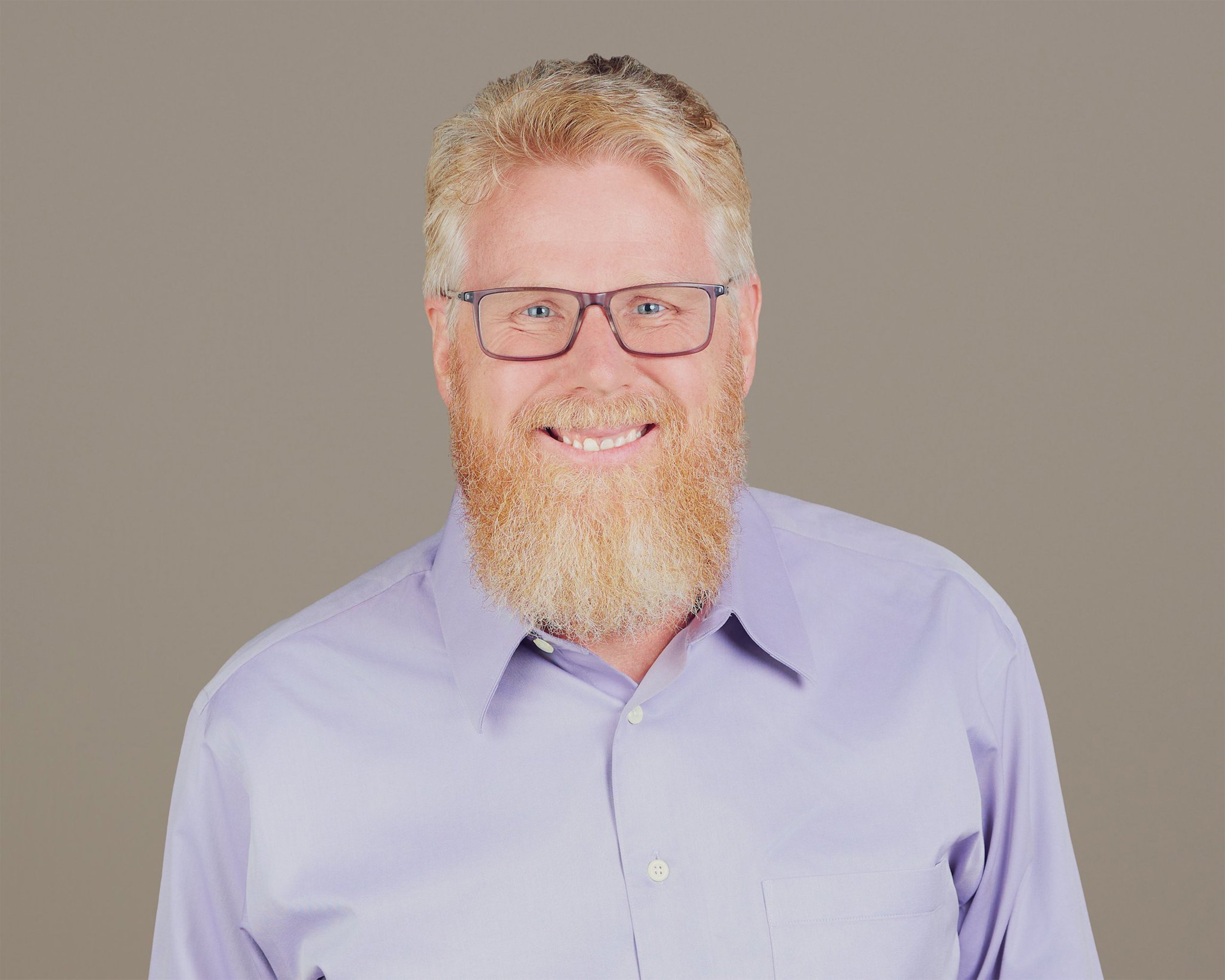 A man with glasses and a red beard smiling, wearing a light purple shirt, against a plain background.