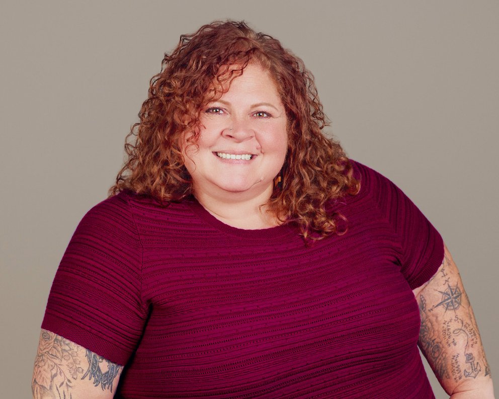 A woman with curly red hair smiling in a maroon top against a plain gray background.