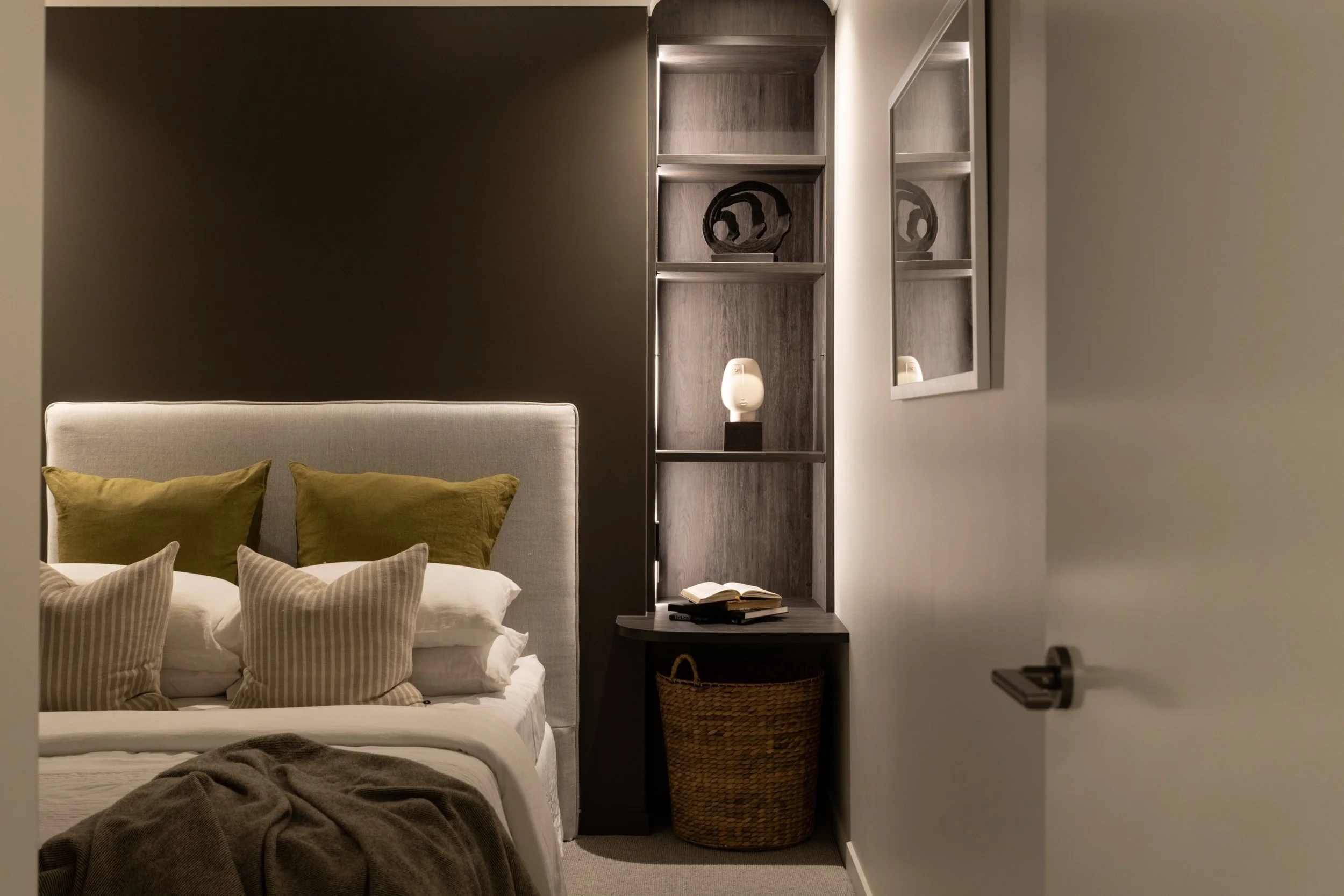 Modern bedroom with gray upholstered headboard, green and striped cushions, gray blanket, and built-in shelving with decorative items and a lamp, alongside a wicker basket and open book on a nightstand.