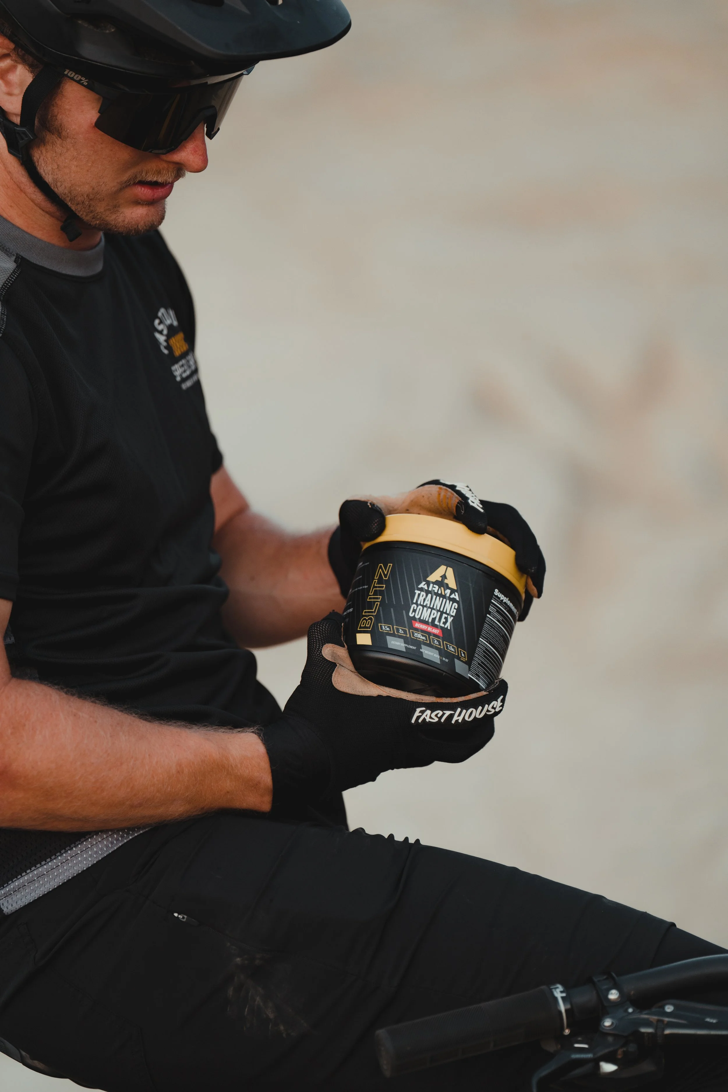 A man wearing a helmet, sunglasses, and asg shirt holds a container of BLITZ training complex supplement while sitting on a bicycle.