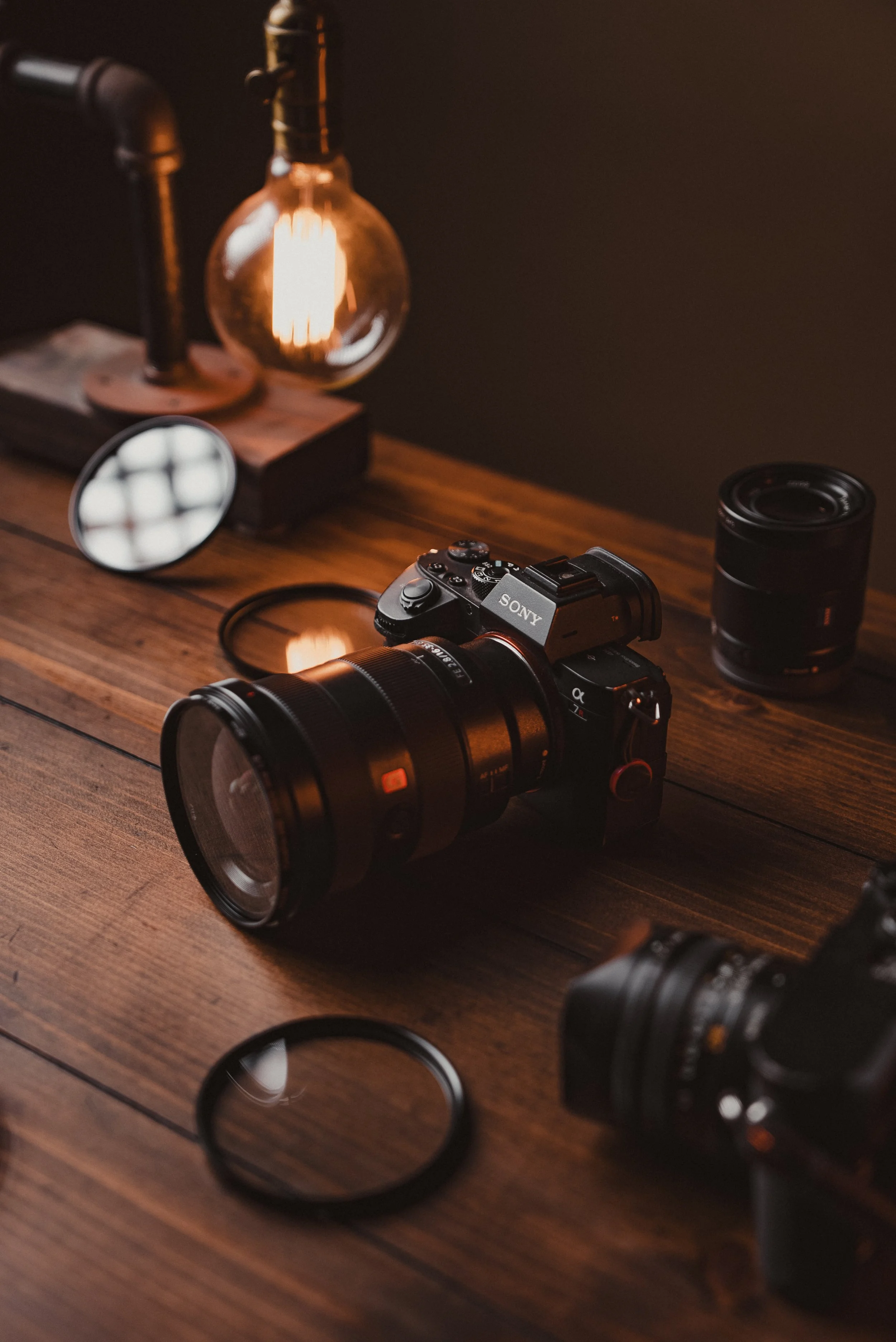 Photography setup with a Sony camera, extra lens, circular filter, and warm ambient lighting on a wooden table.