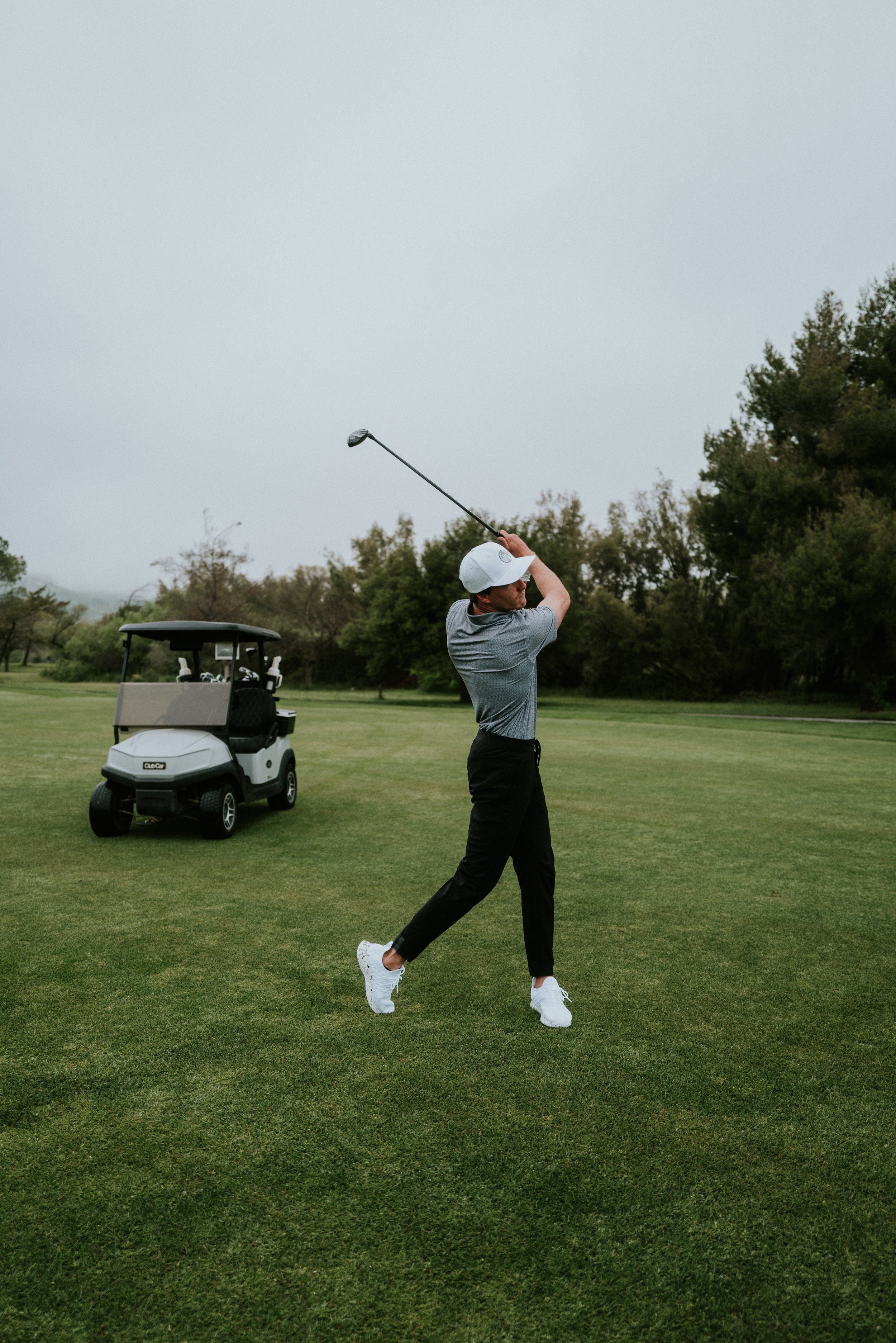 A man playing golf on a golf course, swinging a club with a golf cart nearby, surrounded by trees and grass.