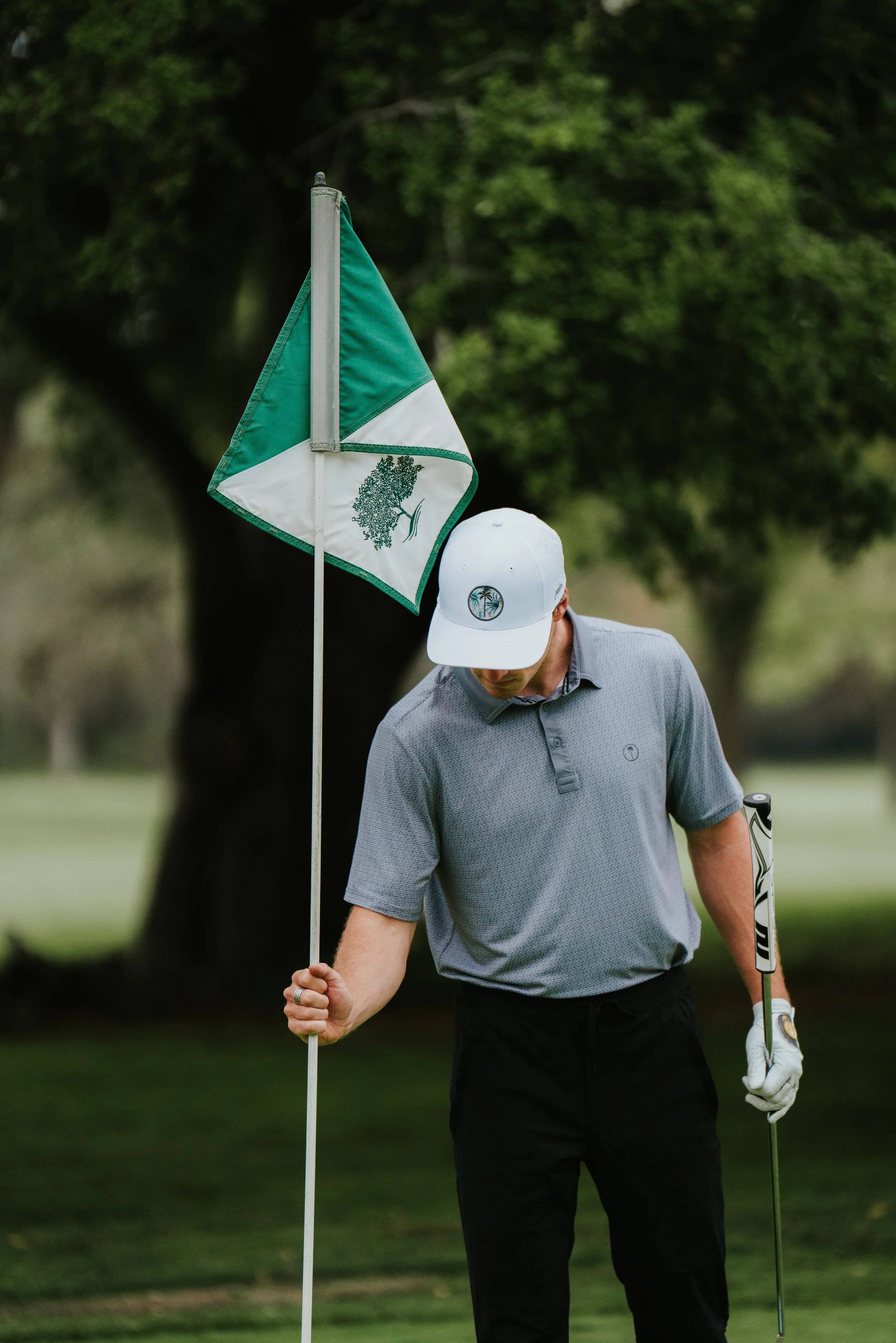 A golfer stands on a golf course holding a flagstick with a green and white flag, with trees and grass in the background.