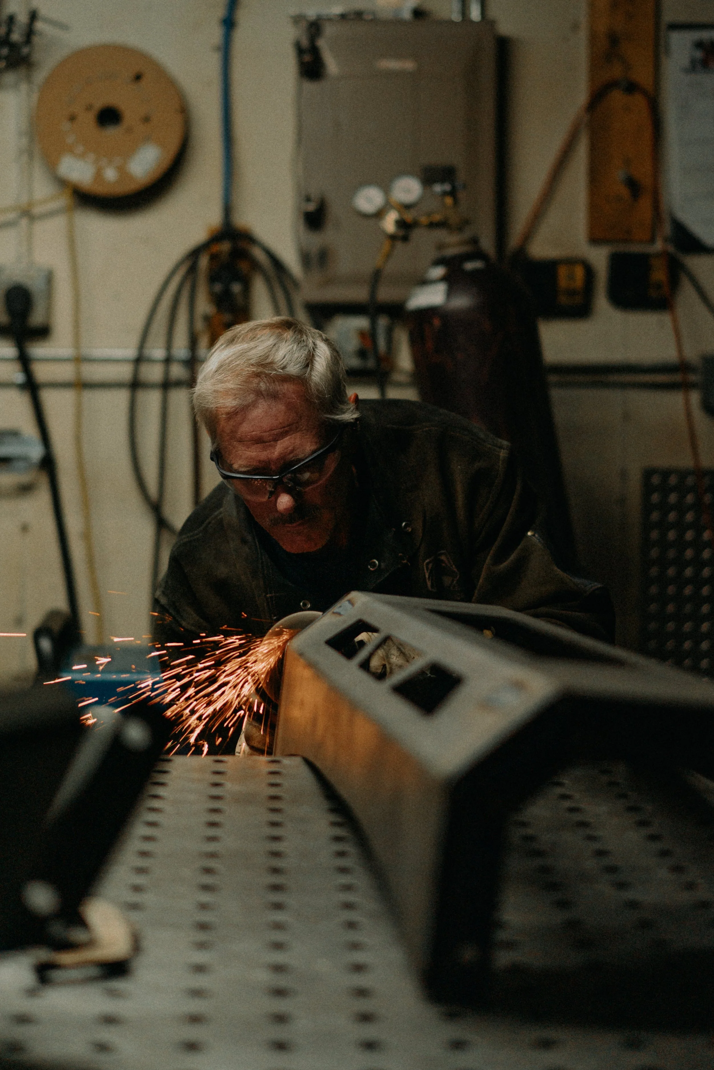 A man wearing protective glasses is welding metal on a workbench in a workshop, with sparks flying from the welding process.