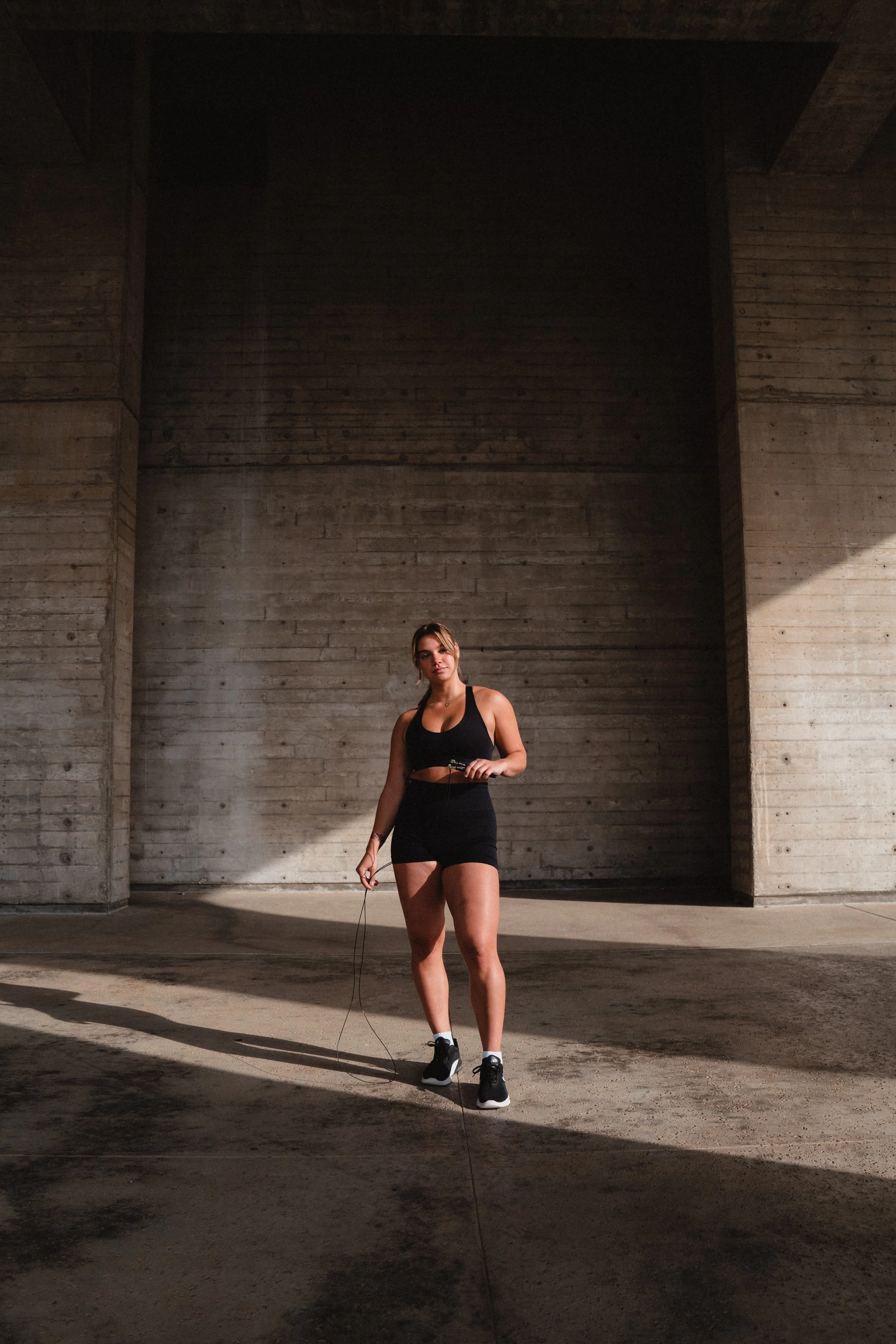 A young woman in workout attire standing in a concrete space, holding a jump rope, with sunlight casting shadows on the ground.