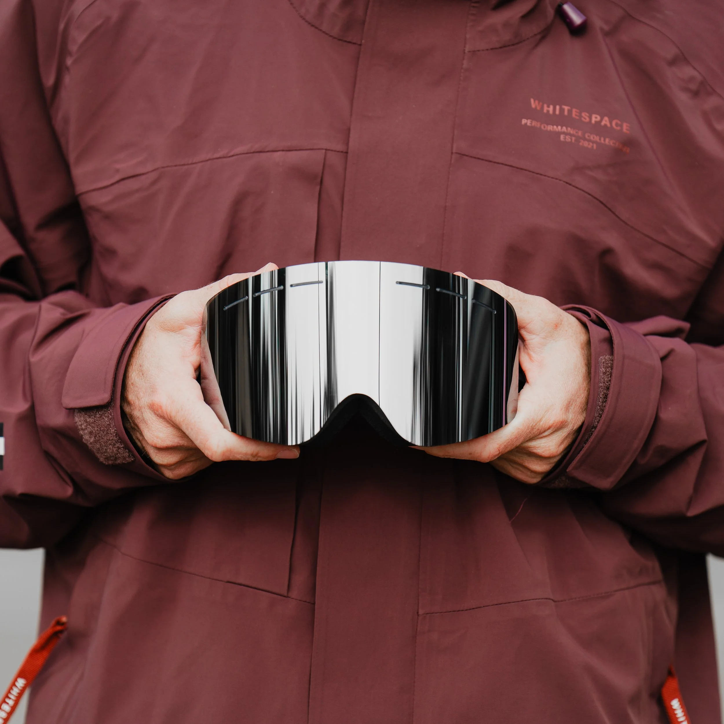 Person wearing a maroon jacket holding a reflective ski or snowboard goggle.