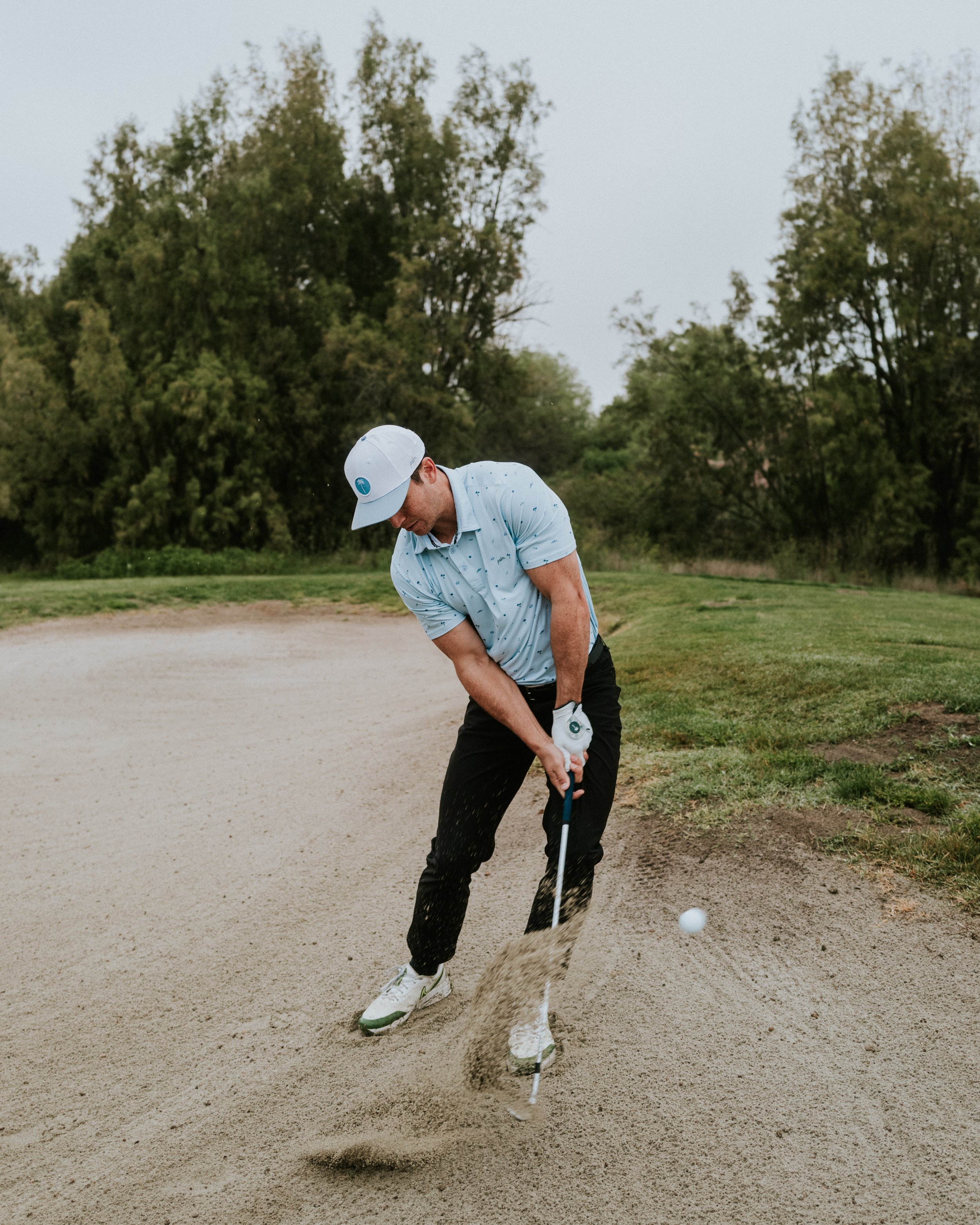 A man in a light blue shirt and cap is hitting a golf ball out of a sand trap on a golf course, with trees in the background.