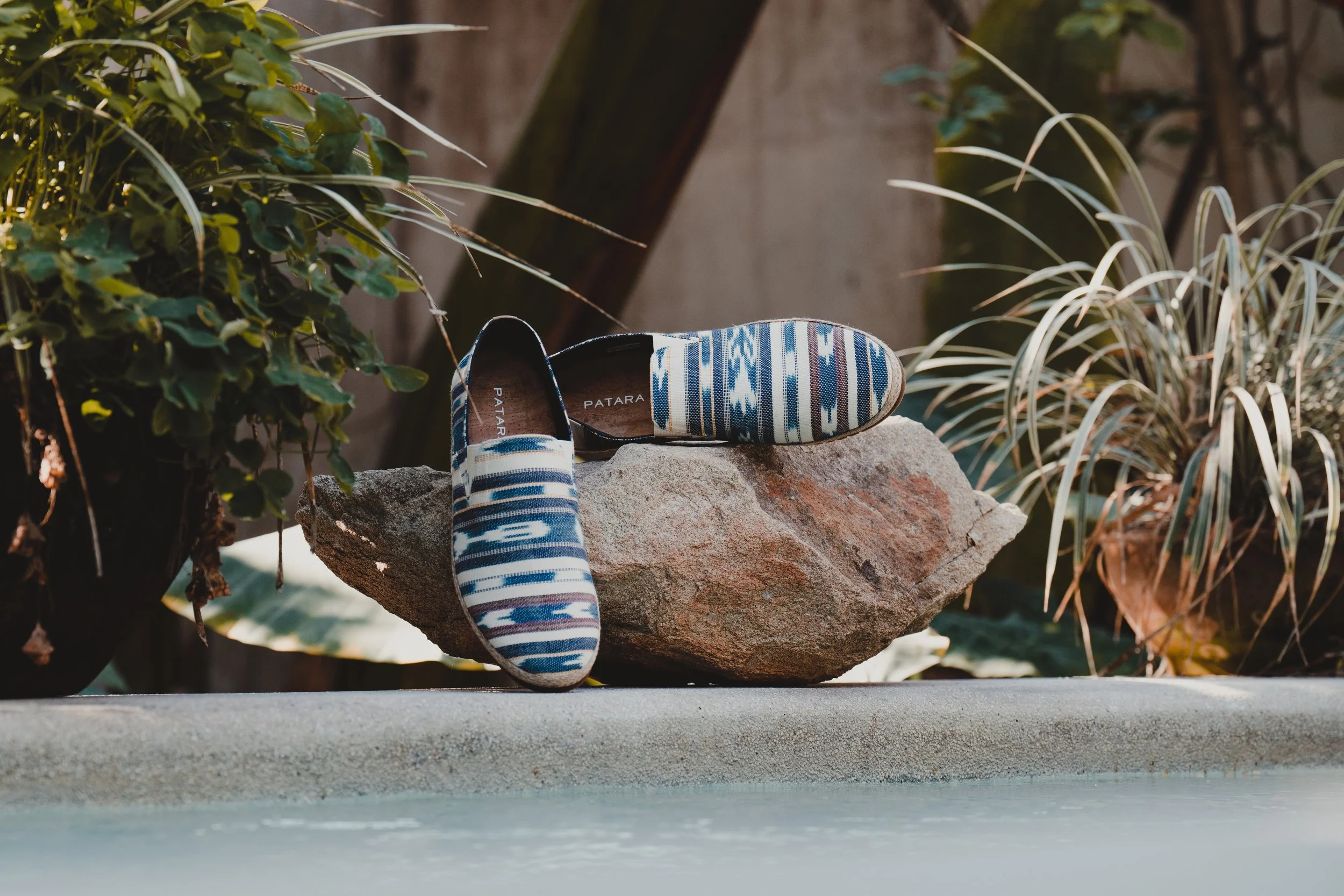 A pair of patterned blue and white slip-on shoes resting on a large rock, with greenery and plants in the background.