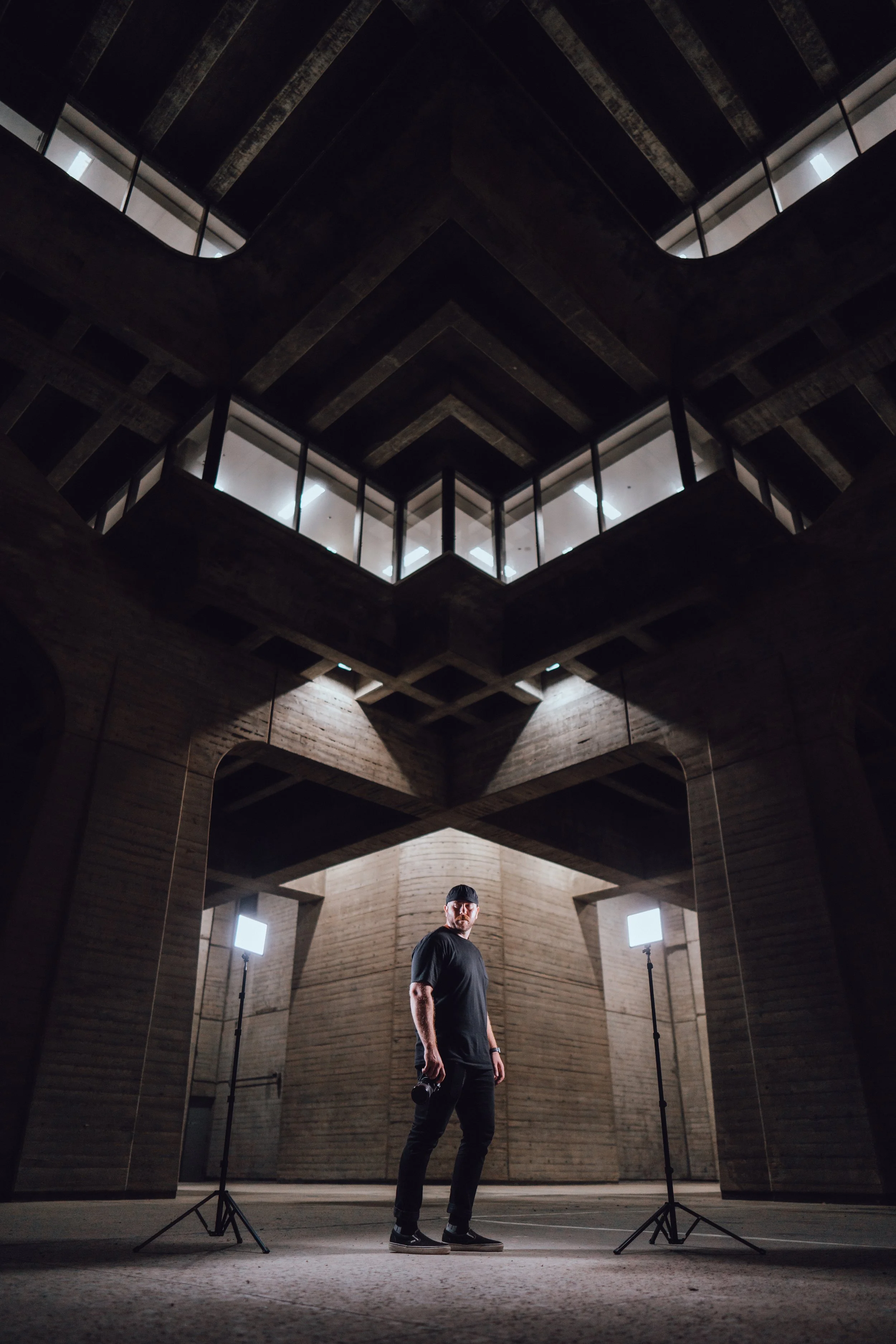 A man in black clothing and a black cap standing in a modern, industrial-style interior with exposed brick walls, illuminated by two bright studio lights.