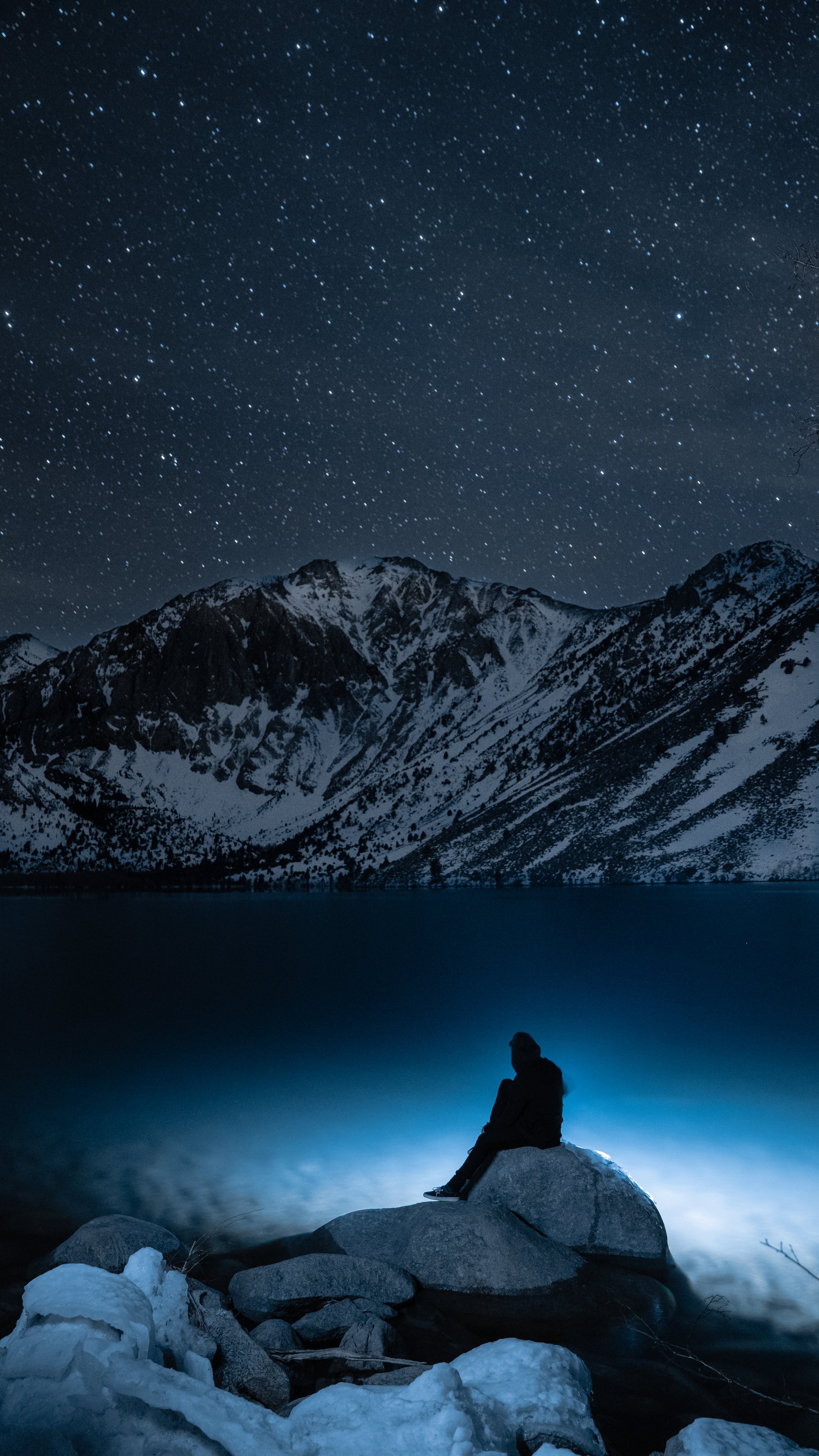 Person sitting on a rock by a lake at night, with snow-covered mountains in the background and a starry sky overhead.
