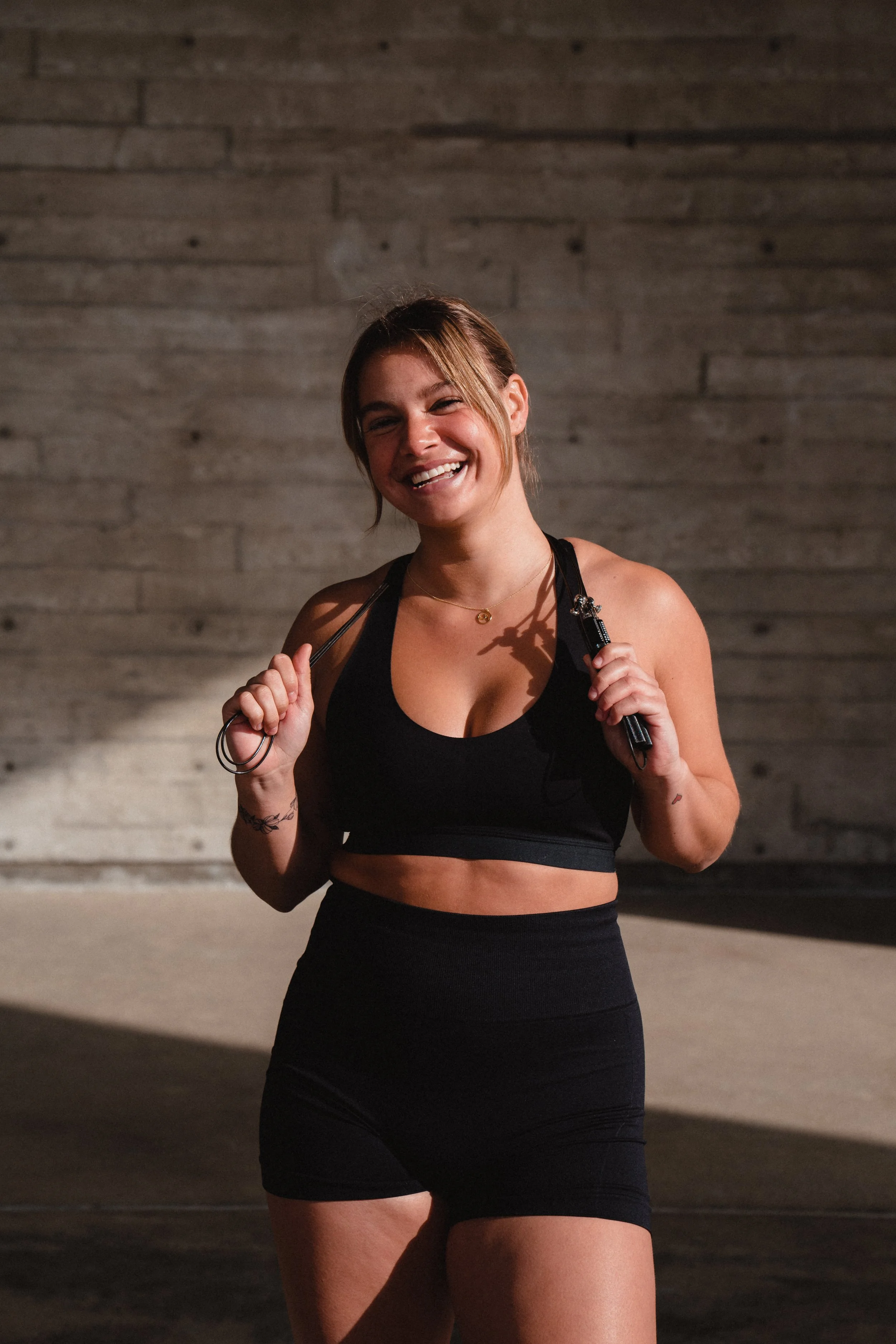 A woman in athletic wear smiling, holding a jump rope over her shoulders, standing in front of a wooden wall.