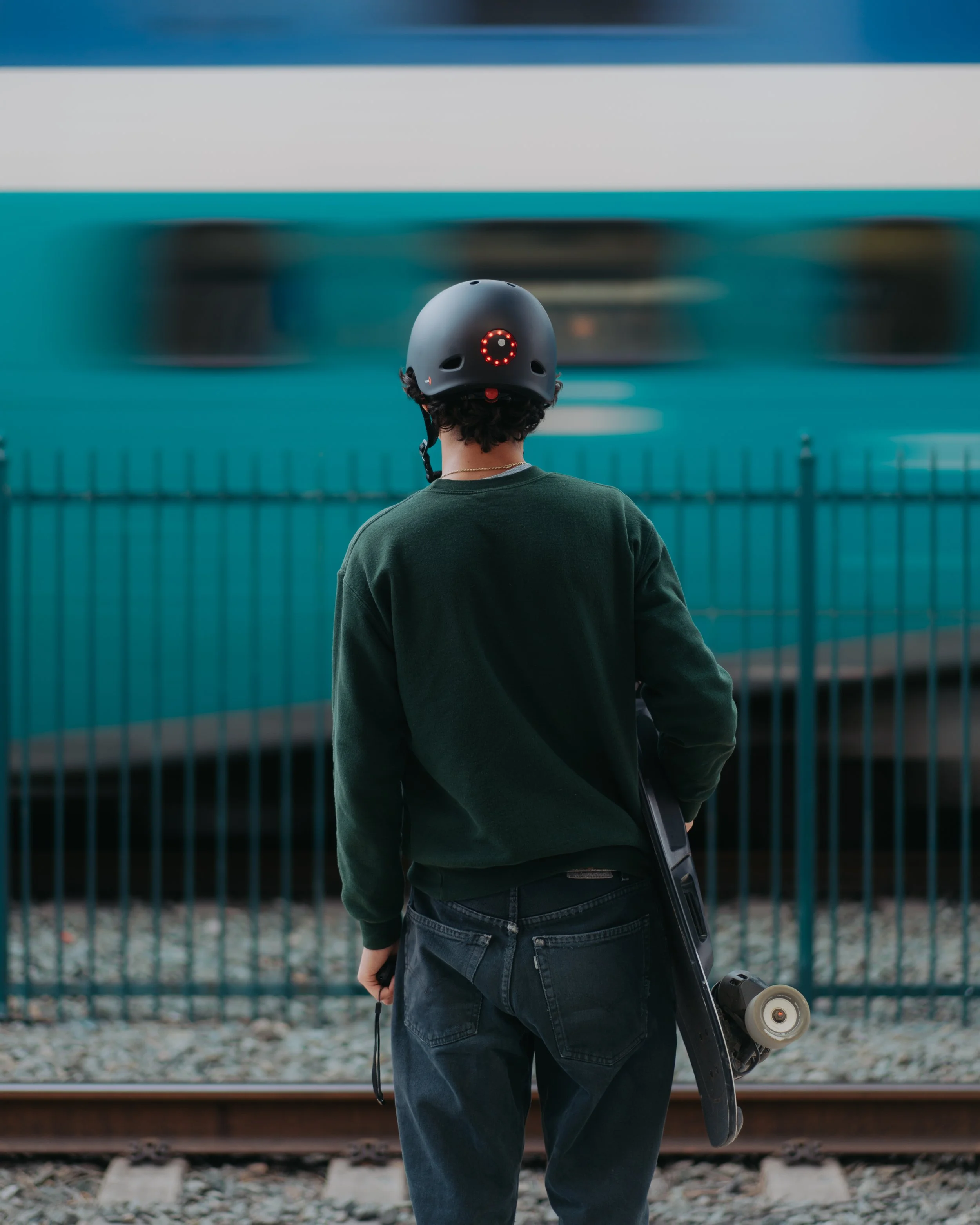 Person wearing a helmet and dark green sweatshirt standing on a train platform, holding a skateboard with a train passing by in the background.