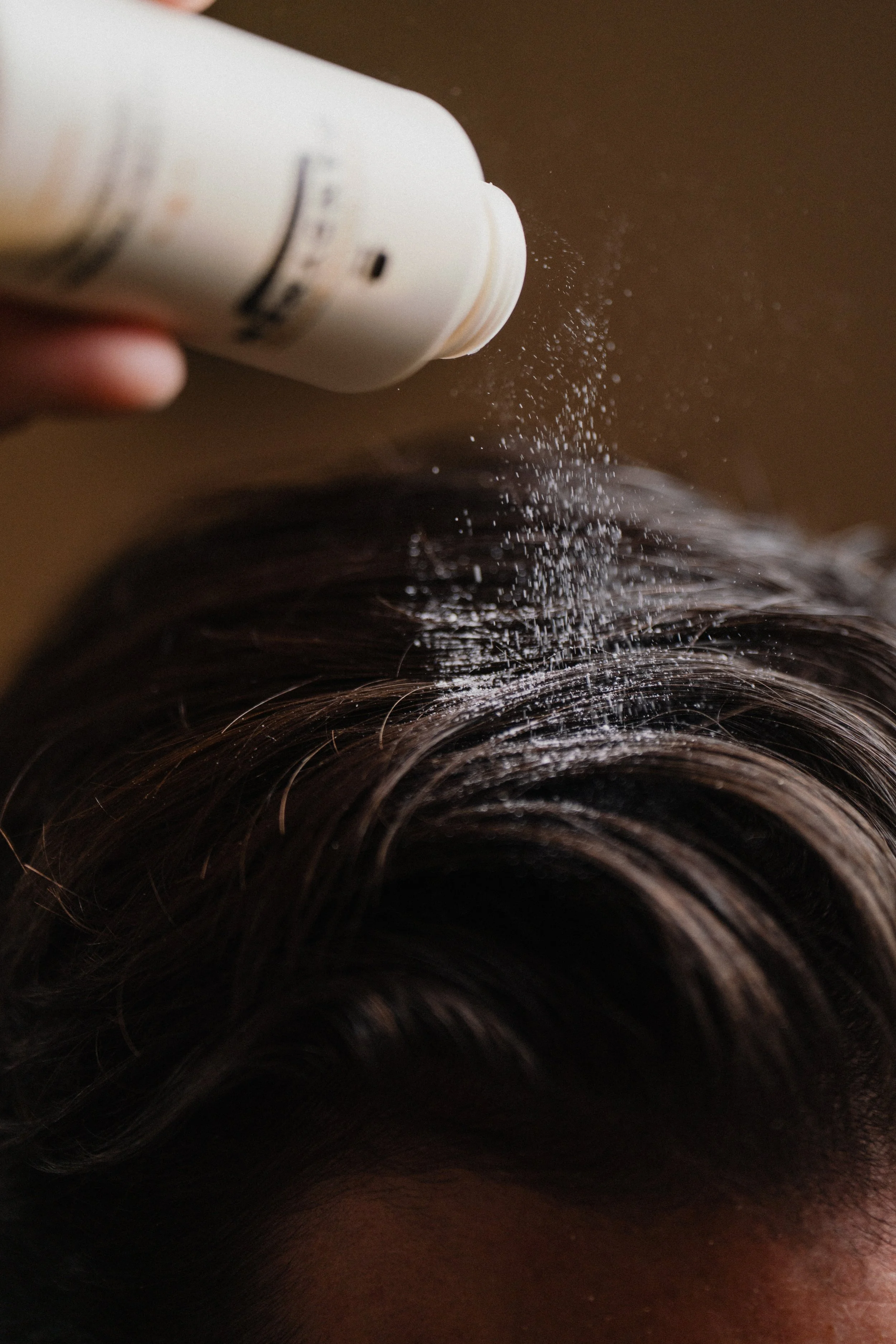 A person sprinkling white powder, likely hair product or dry shampoo, onto dark brown, wavy hair.