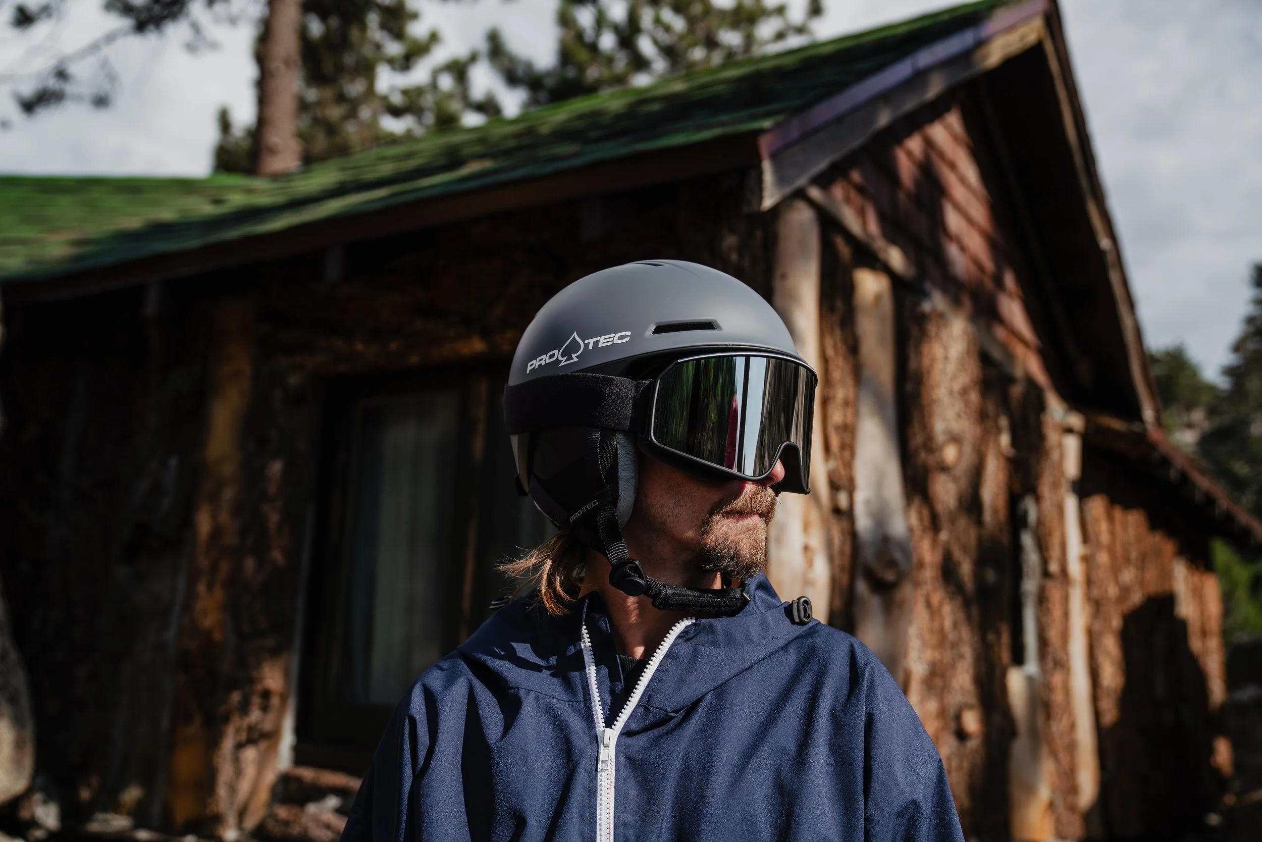 A man wearing a Pro Tec motorcycle helmet with goggles and a navy jacket stands outdoors near a rustic wooden house.