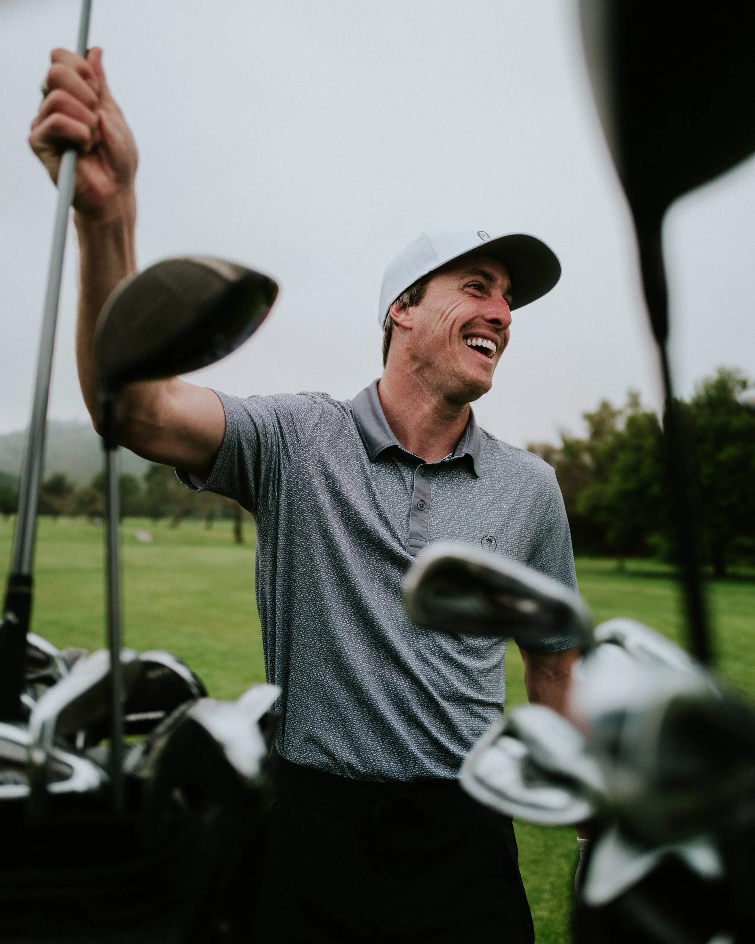 A man smiling on a golf course, holding a golf club with golf bags and clubs nearby.