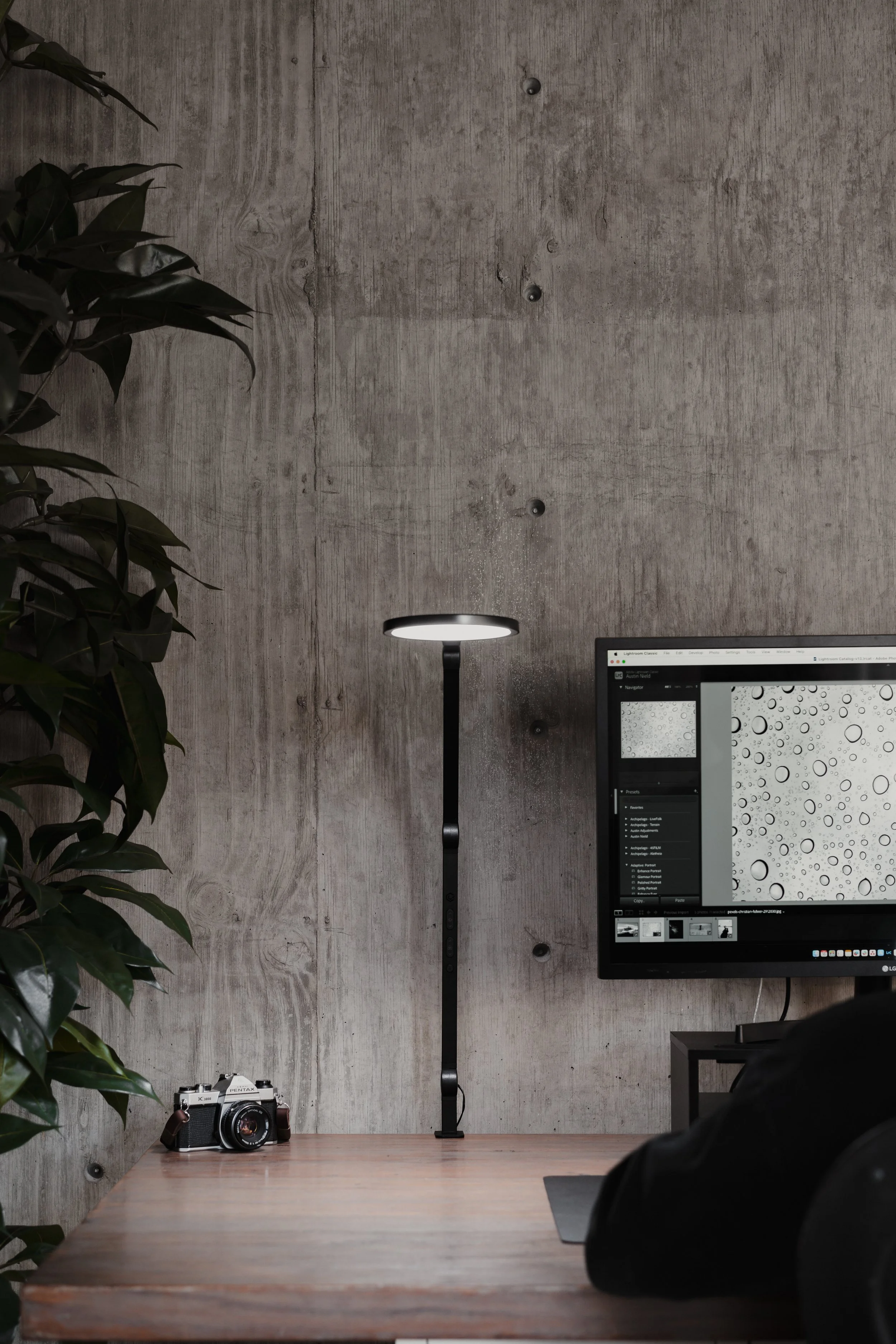 A minimalist workspace with a concrete wall, a desk lamp, a vintage camera on a wooden desk, and a computer monitor displaying droplets on a screen.