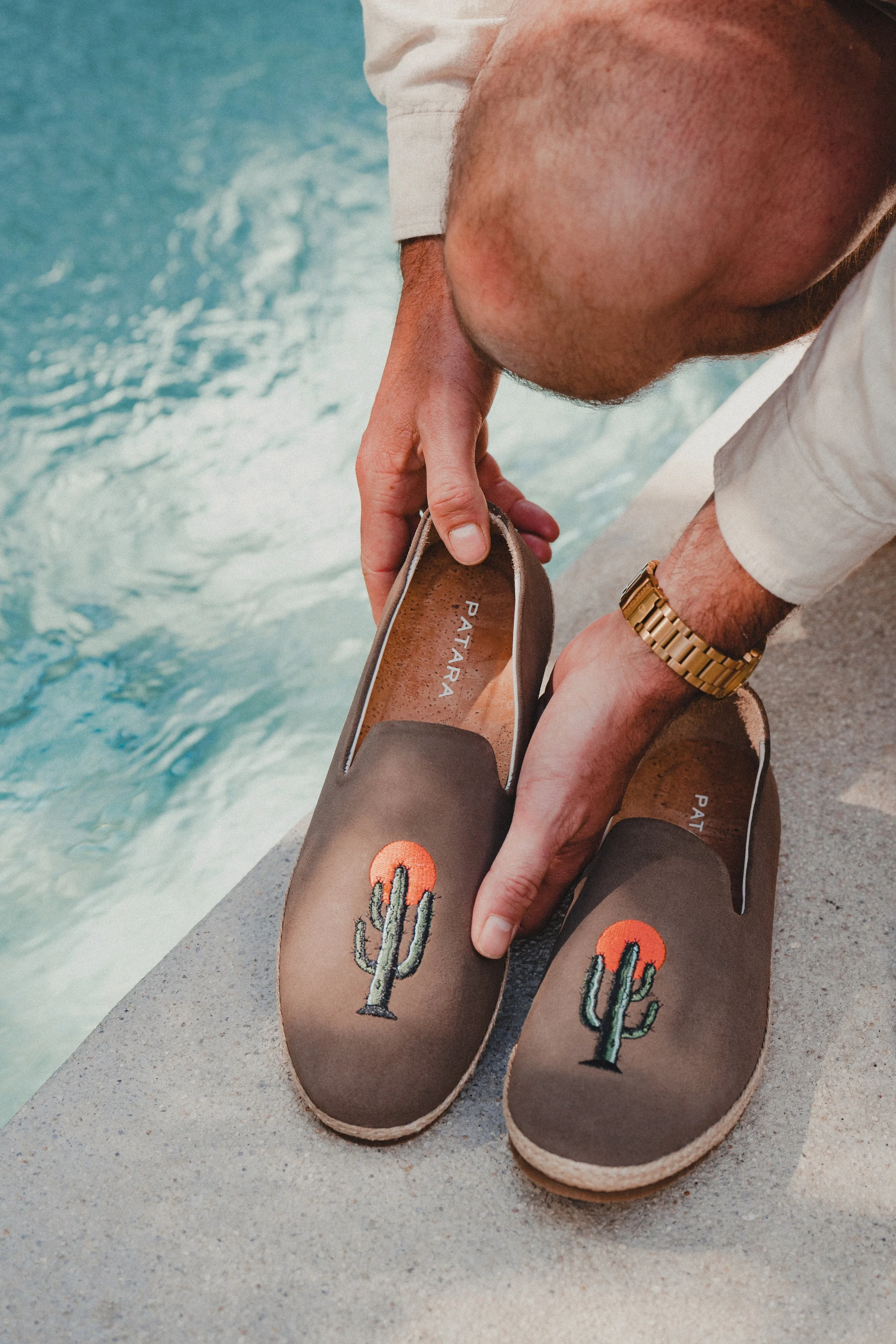 A man wearing a gold watch is placing brown slip-on shoes with cactus and sunset embroidery on a concrete ledge beside a pool.