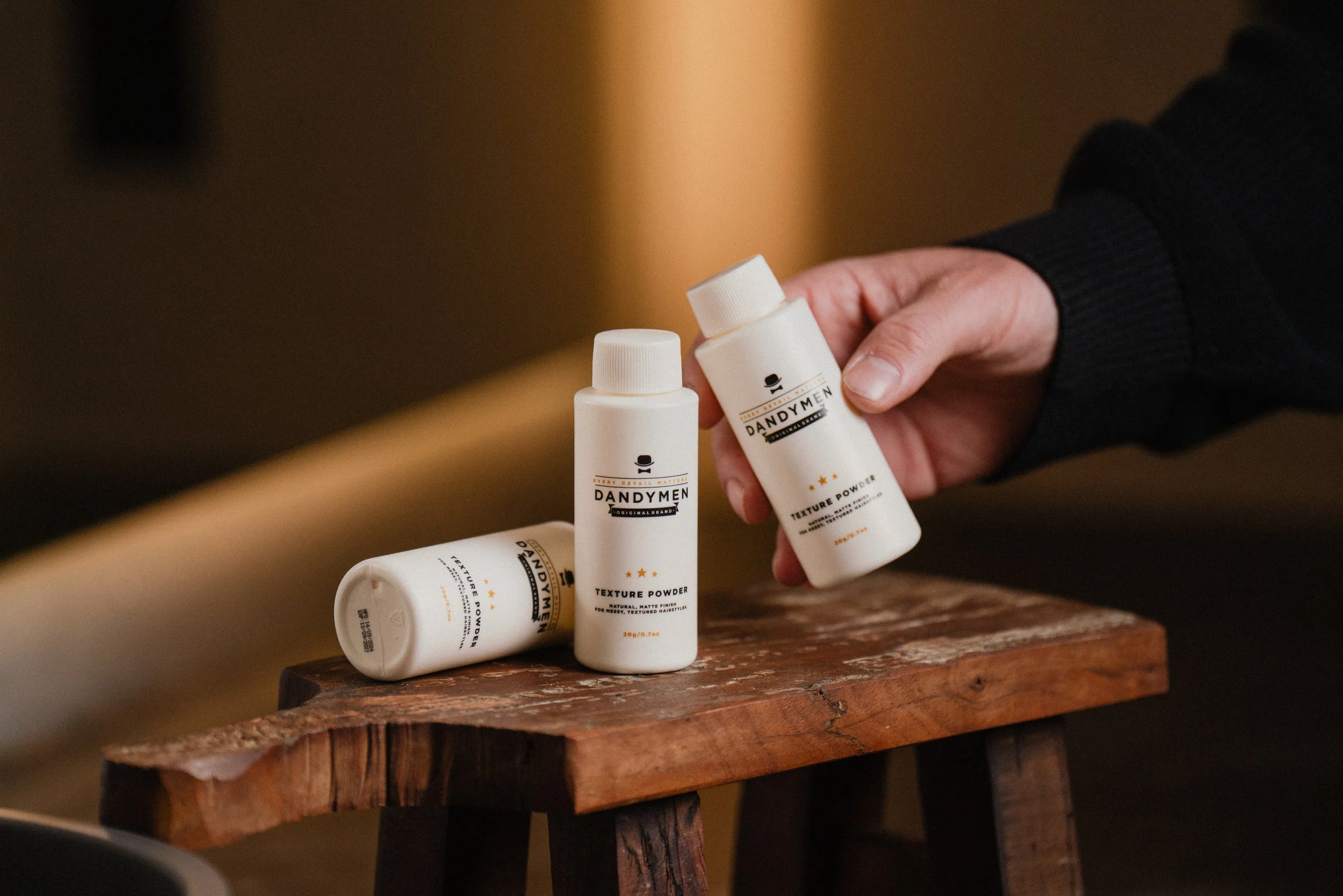 Person holding a white bottle of Dandymen texture powder on a wooden table with two other matching bottles lying down.