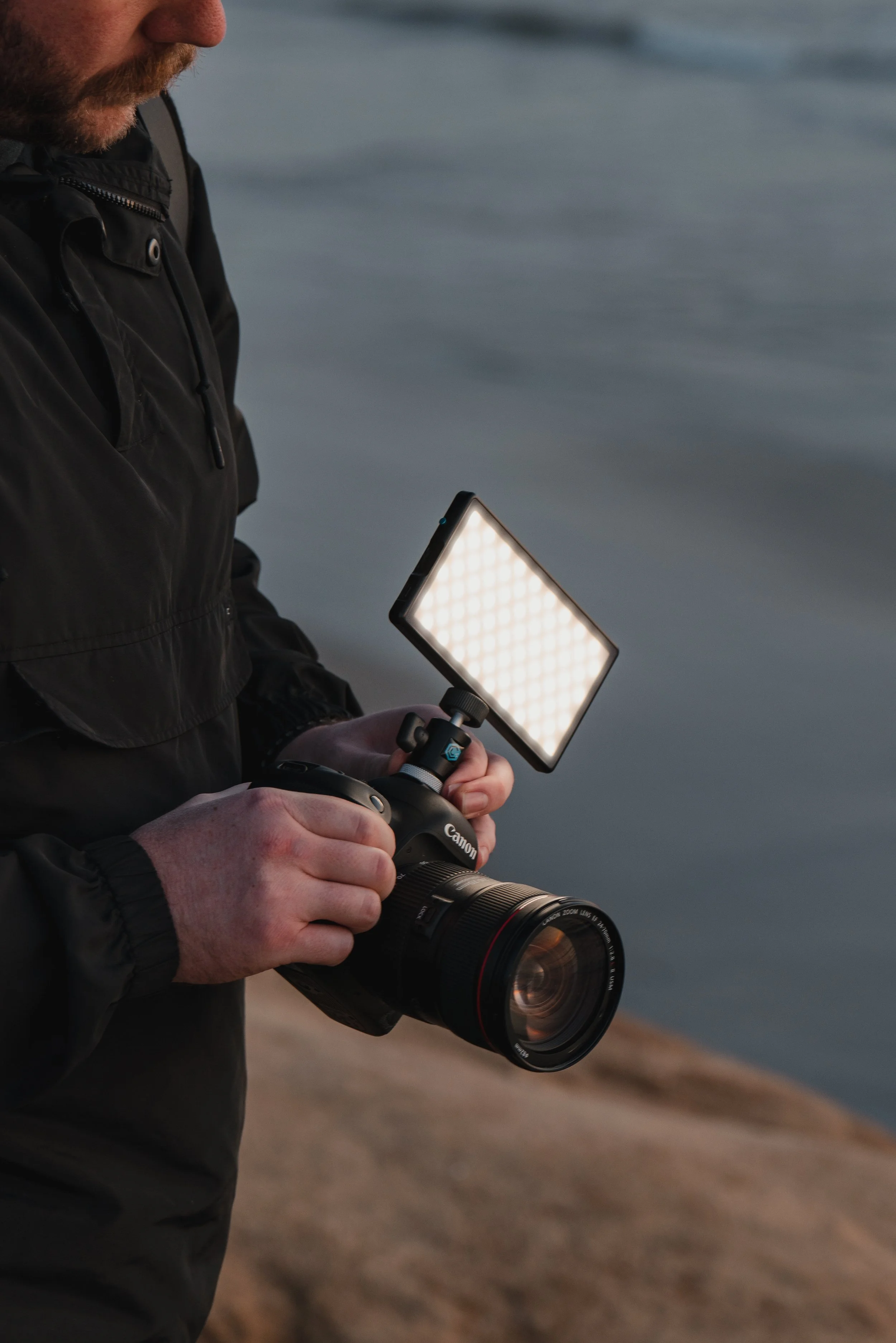 A person holding a Canon camera with an attached LED light panel, near a body of water.