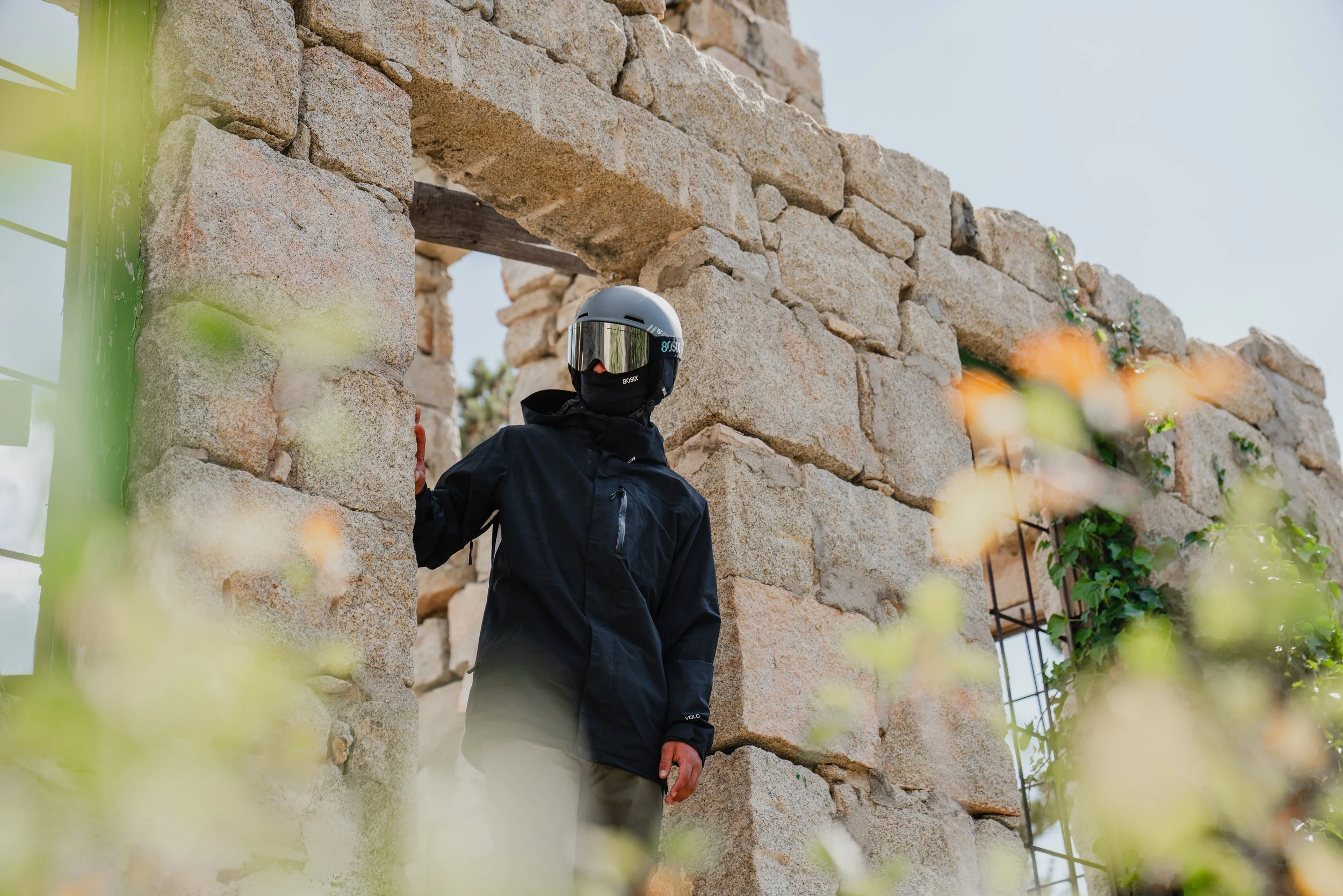 Person wearing a black jacket and a helmet with a visor standing next to a stone wall ruins, surrounded by blurred green plants.