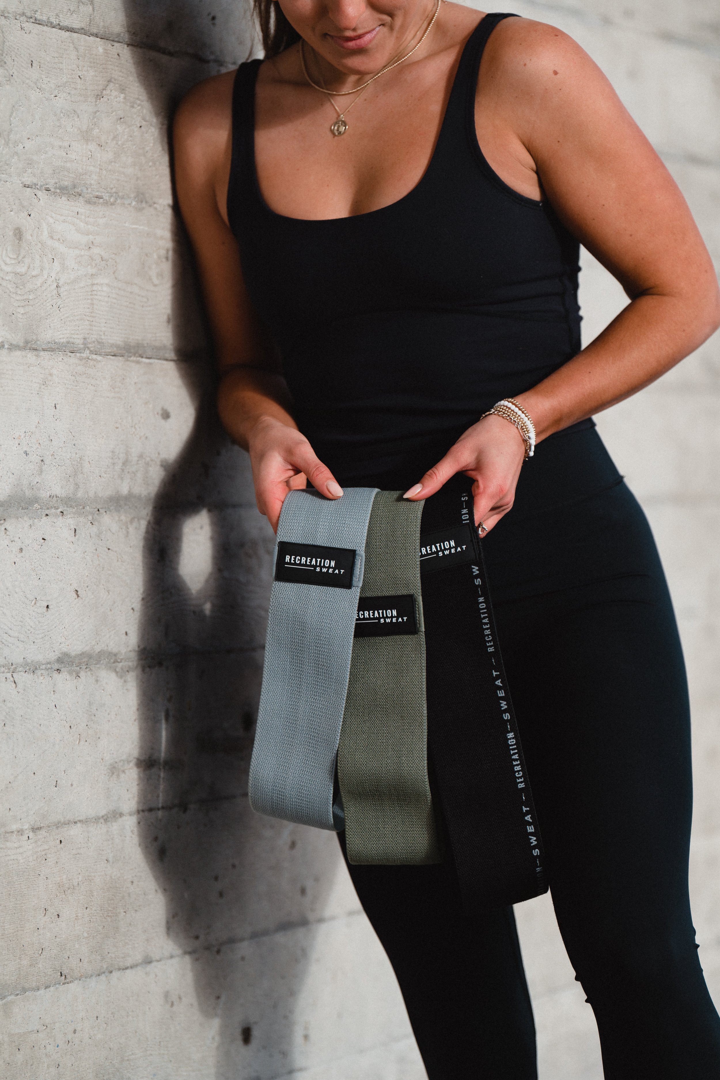 A woman in black athletic clothing stands against a concrete wall, holding three resistance bands labeled with the words "Recreation Sweat."