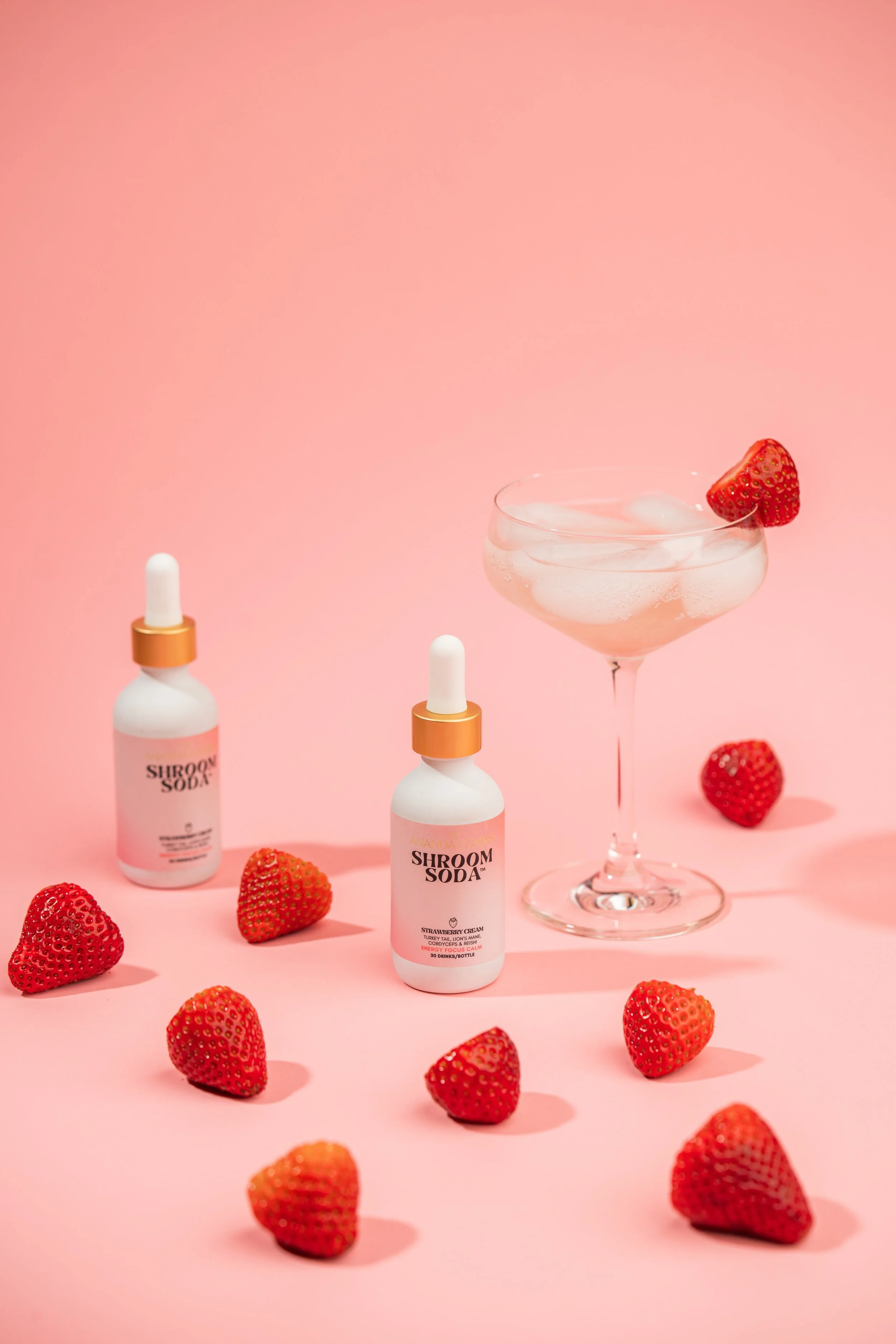 A pink background with two bottles labeled 'Shroom Soda,' a cocktail glass with strawberries and soda inside, and whole strawberries scattered around.