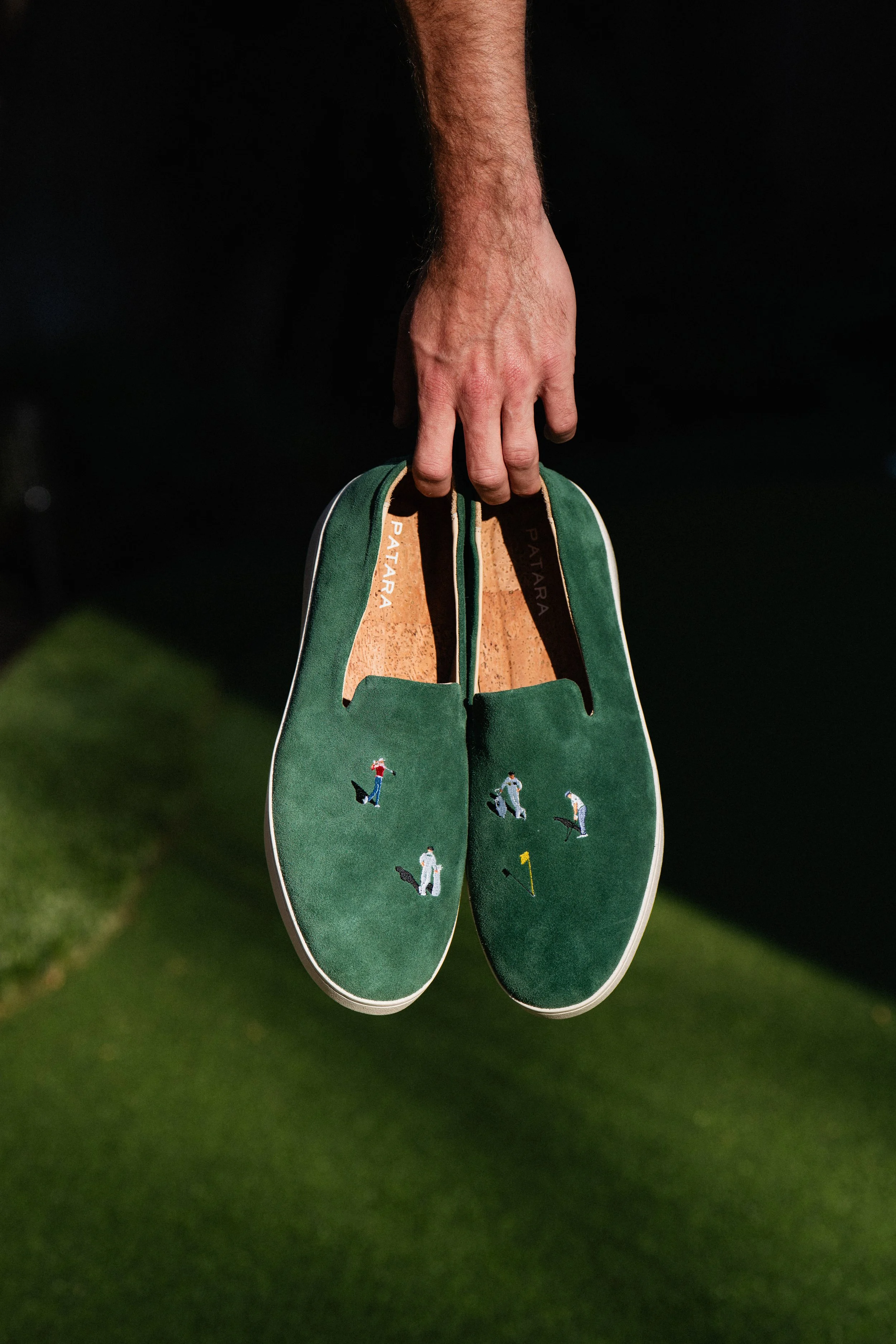 A person's hand holding a pair of green slip-on shoes with embroidered images of people playing golf, with a grassy area in the background.