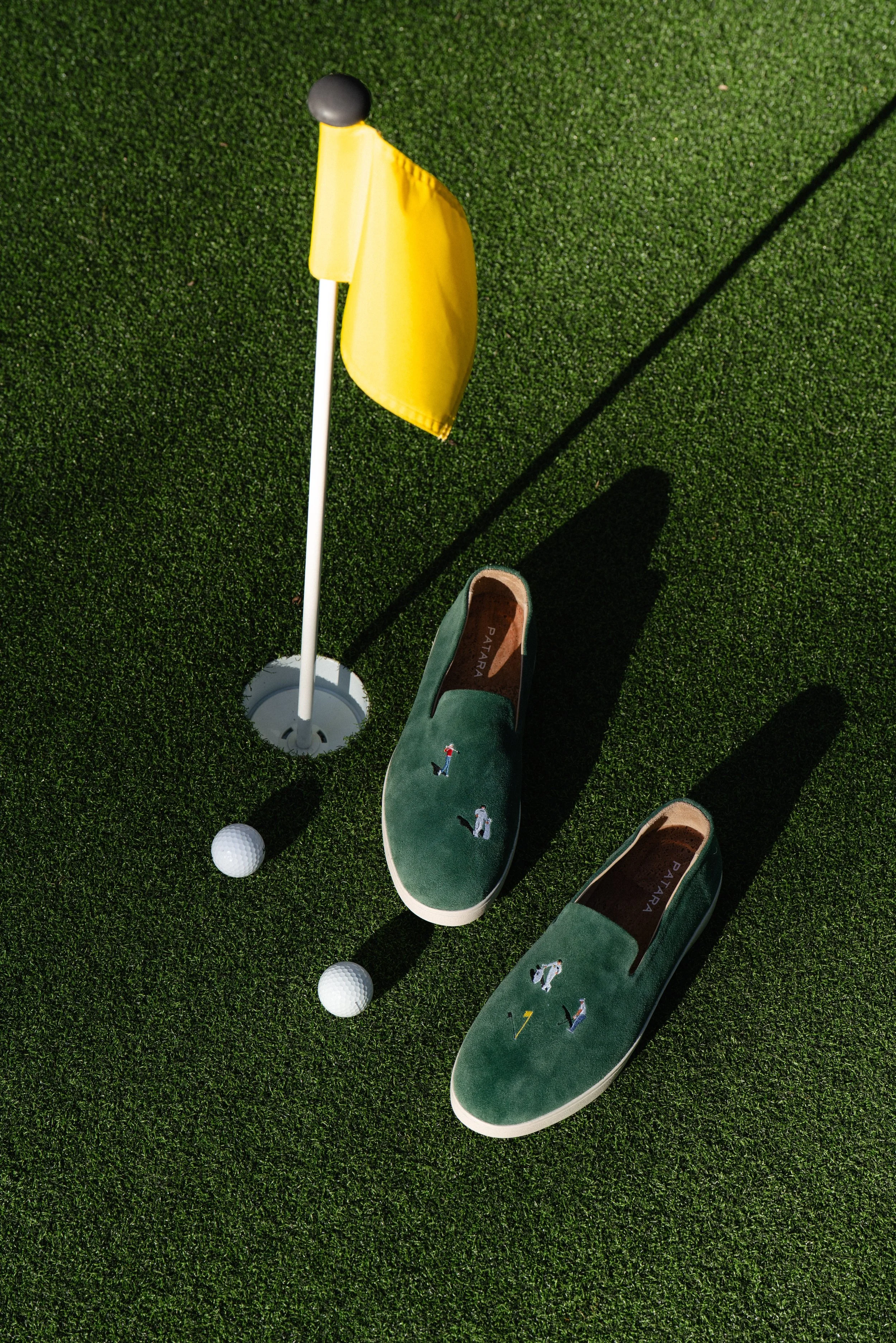 A pair of green slip-on shoes with embroidered golf figures on a putting green, a yellow golf flag, and three golf balls.