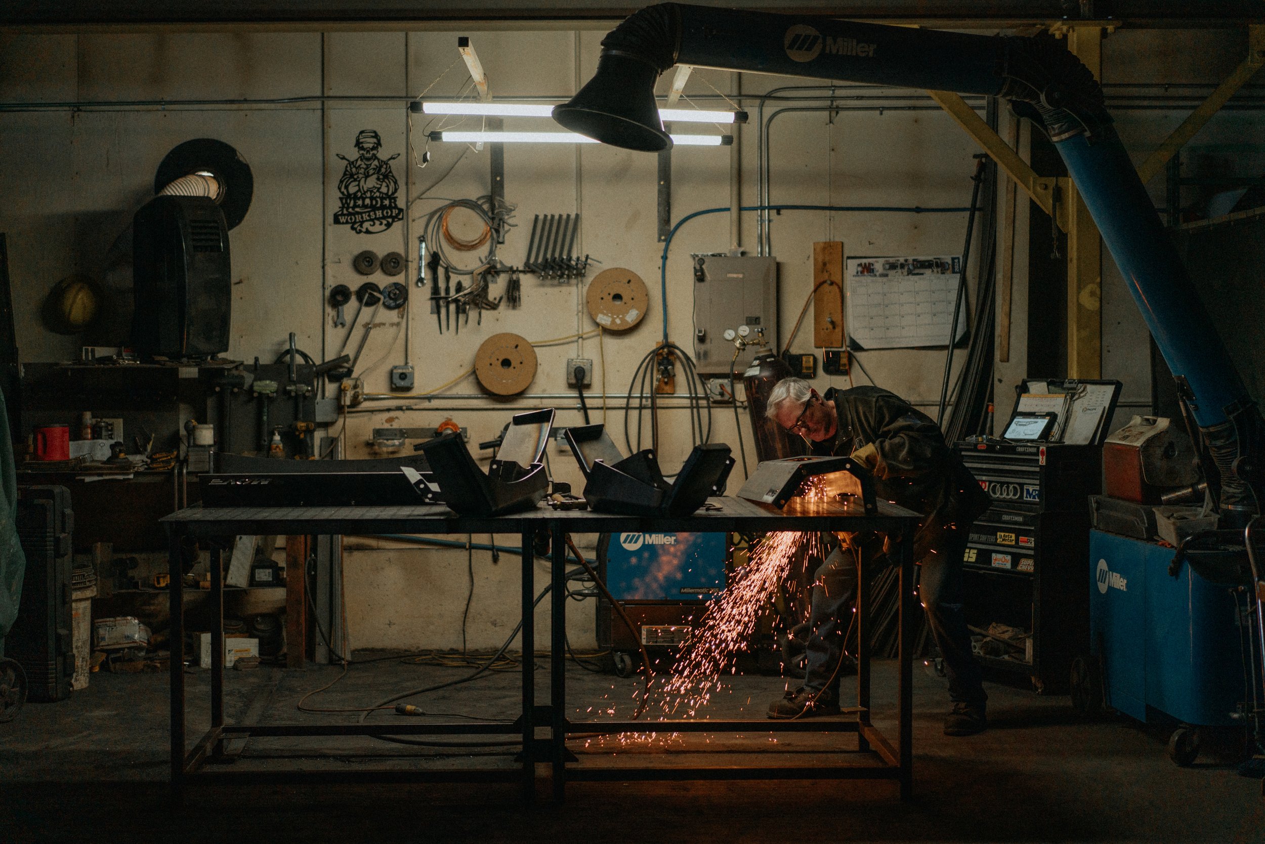A man welding in a workshop, sparks flying as he works at a metal table surrounded by tools and equipment.