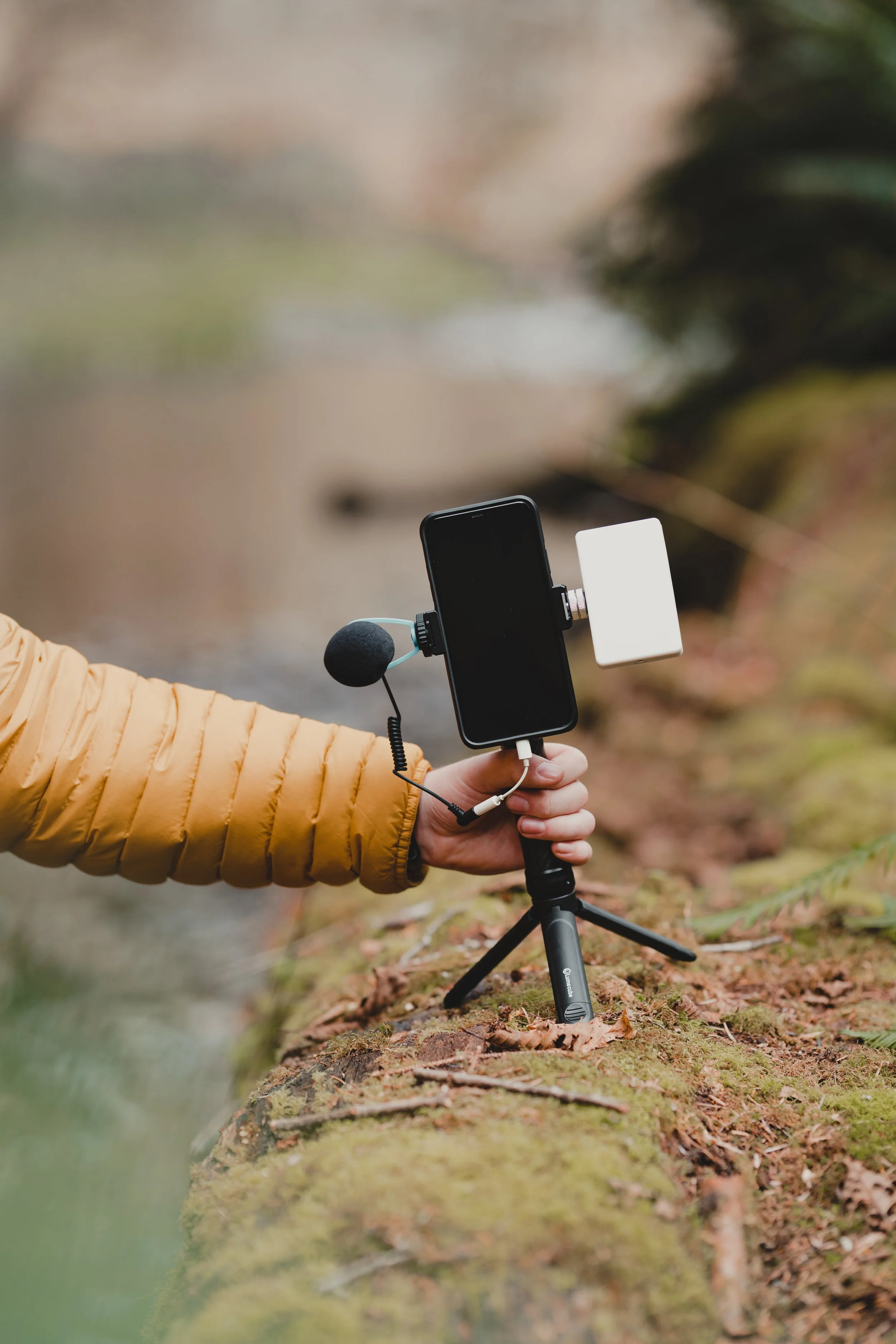 A person in a yellow jacket holds a smartphone with a microphone and a portable power bank attached, set up on a small tripod on a forest floor.