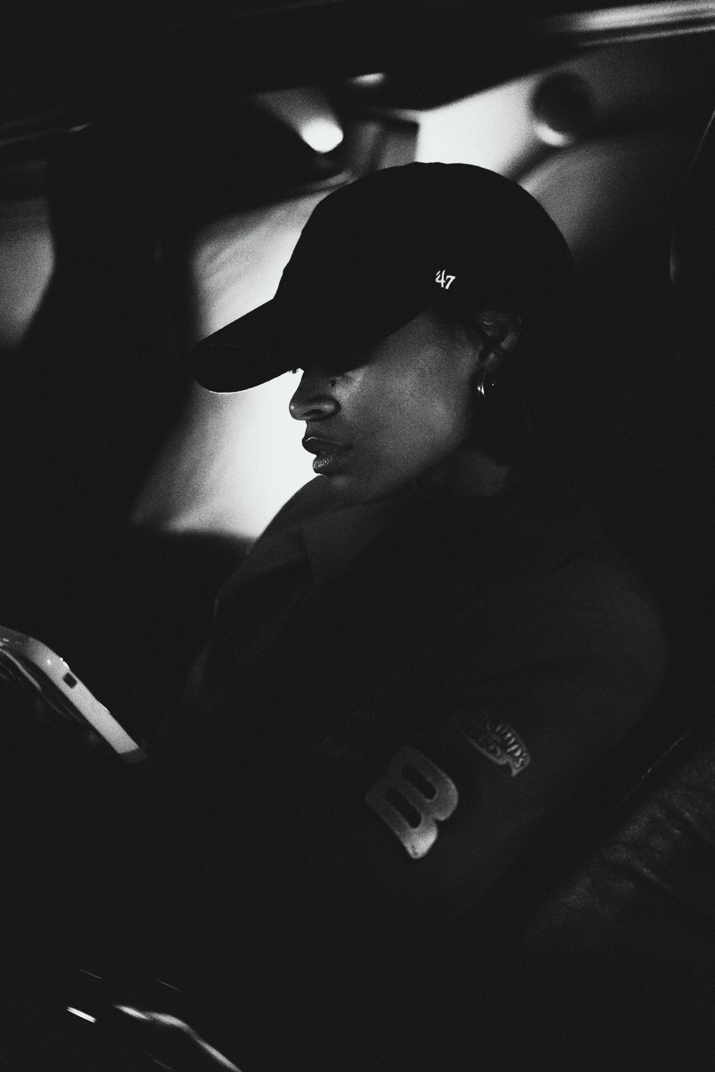 A woman wearing a black baseball cap with the number 47, sitting in a car, looking down at her phone, with a dark and minimal background.