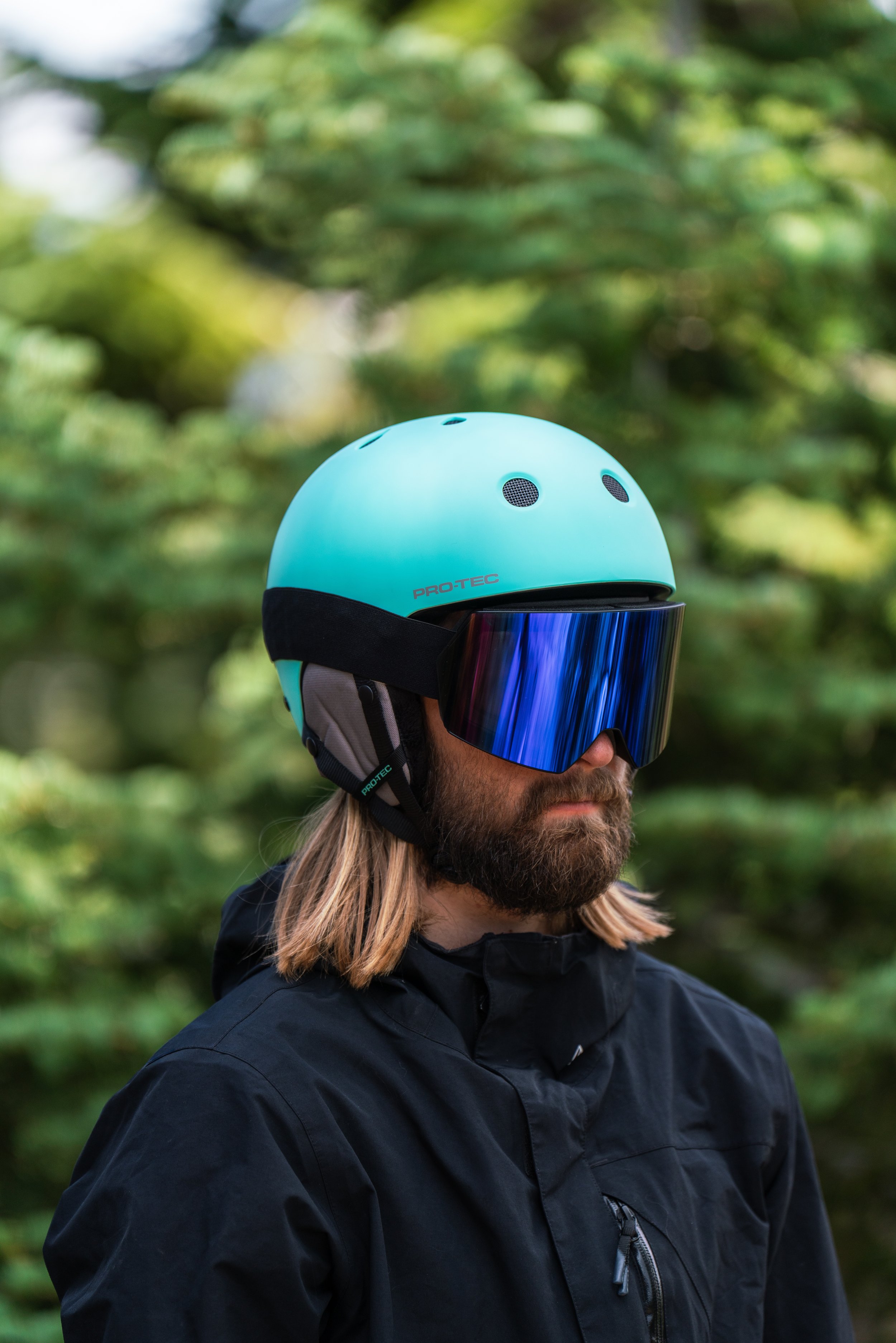 Man with long blond hair and beard wearing a black jacket, a teal helmet, and ski goggles outdoors in a wooded area.