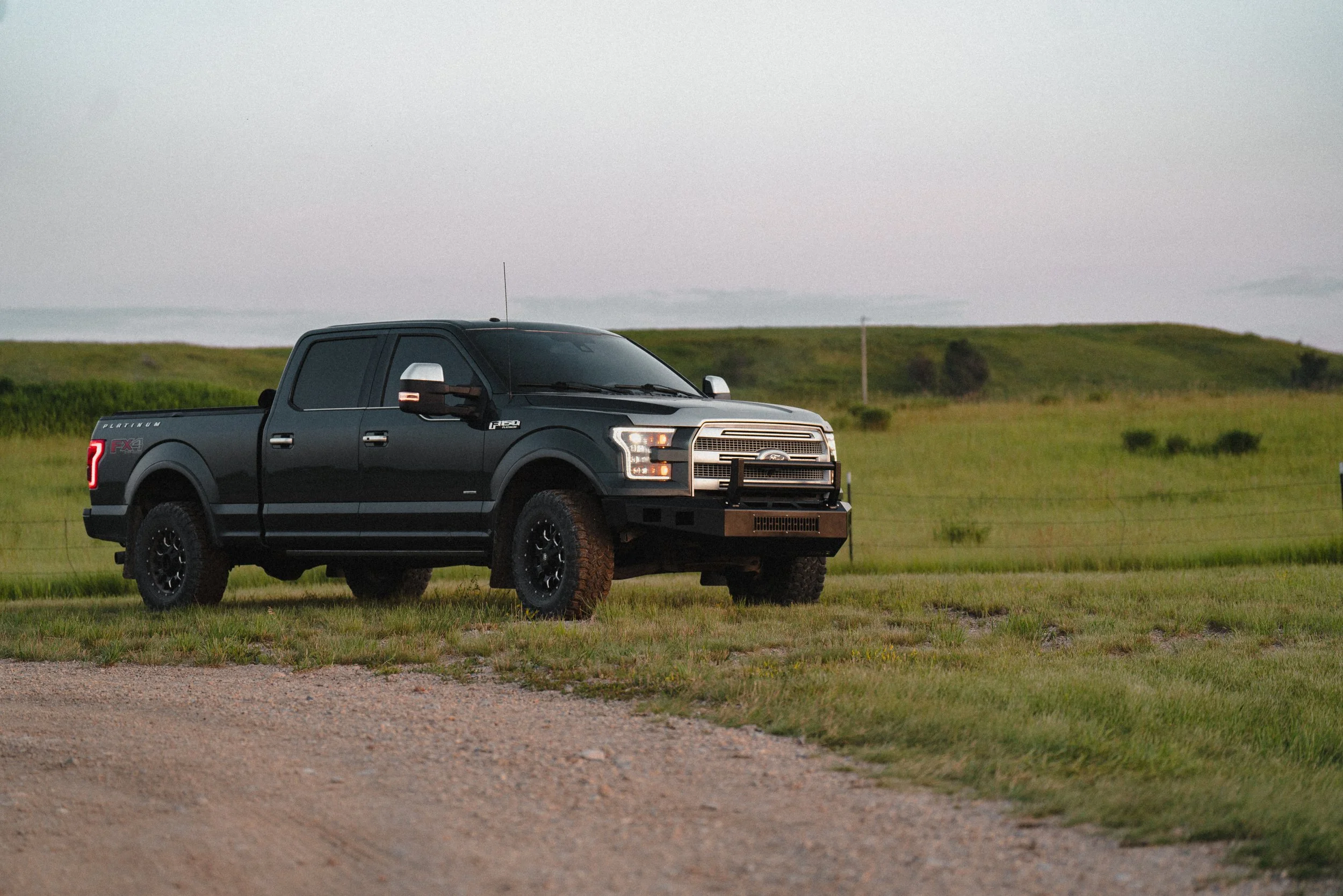 Black Ford F-150 pickup truck parked on a grassy field during daytime with a cloudy sky and rolling hills in the background.
