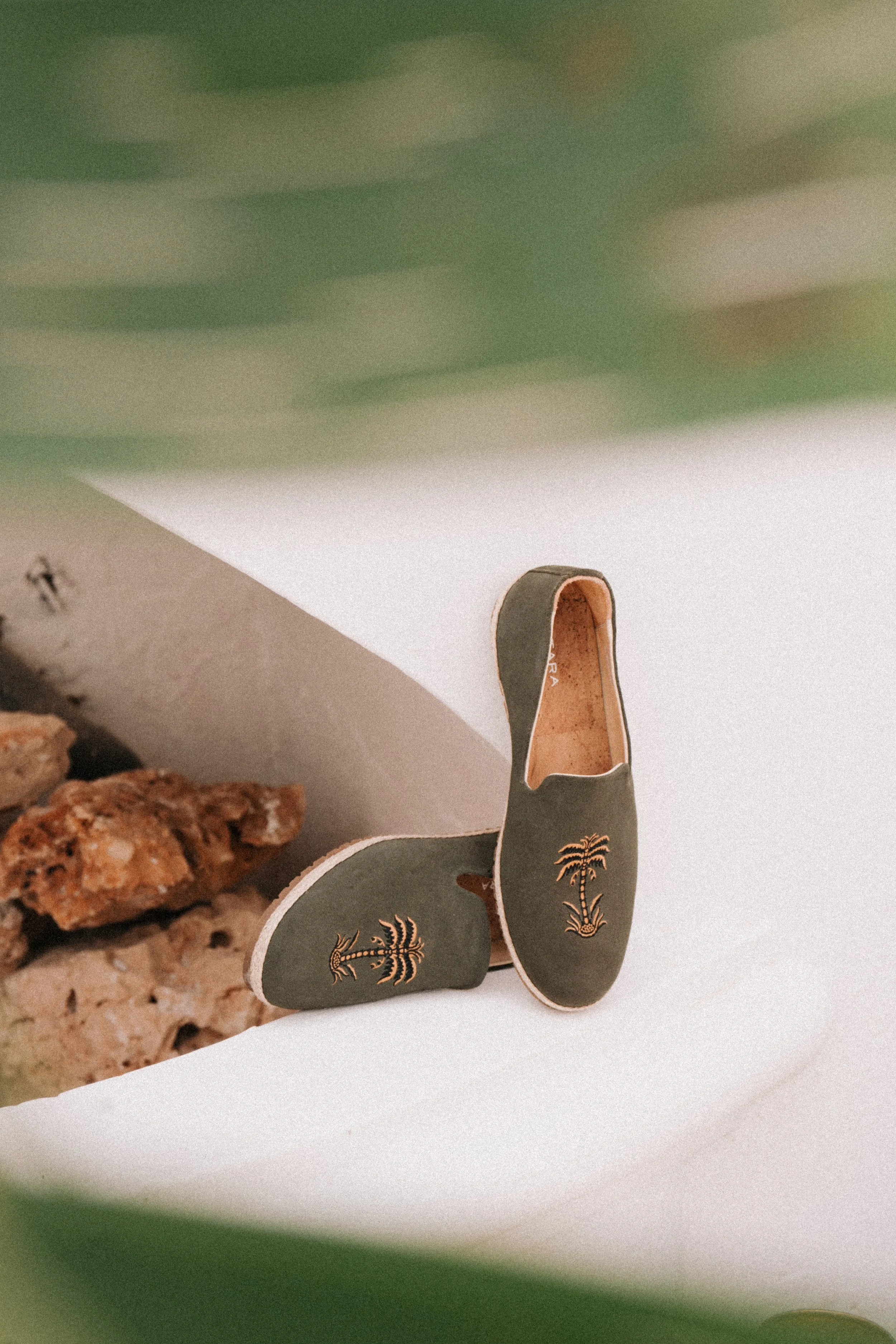 A pair of children's slip-on shoes with palm tree embroidery, positioned on a white surface near a stone area.