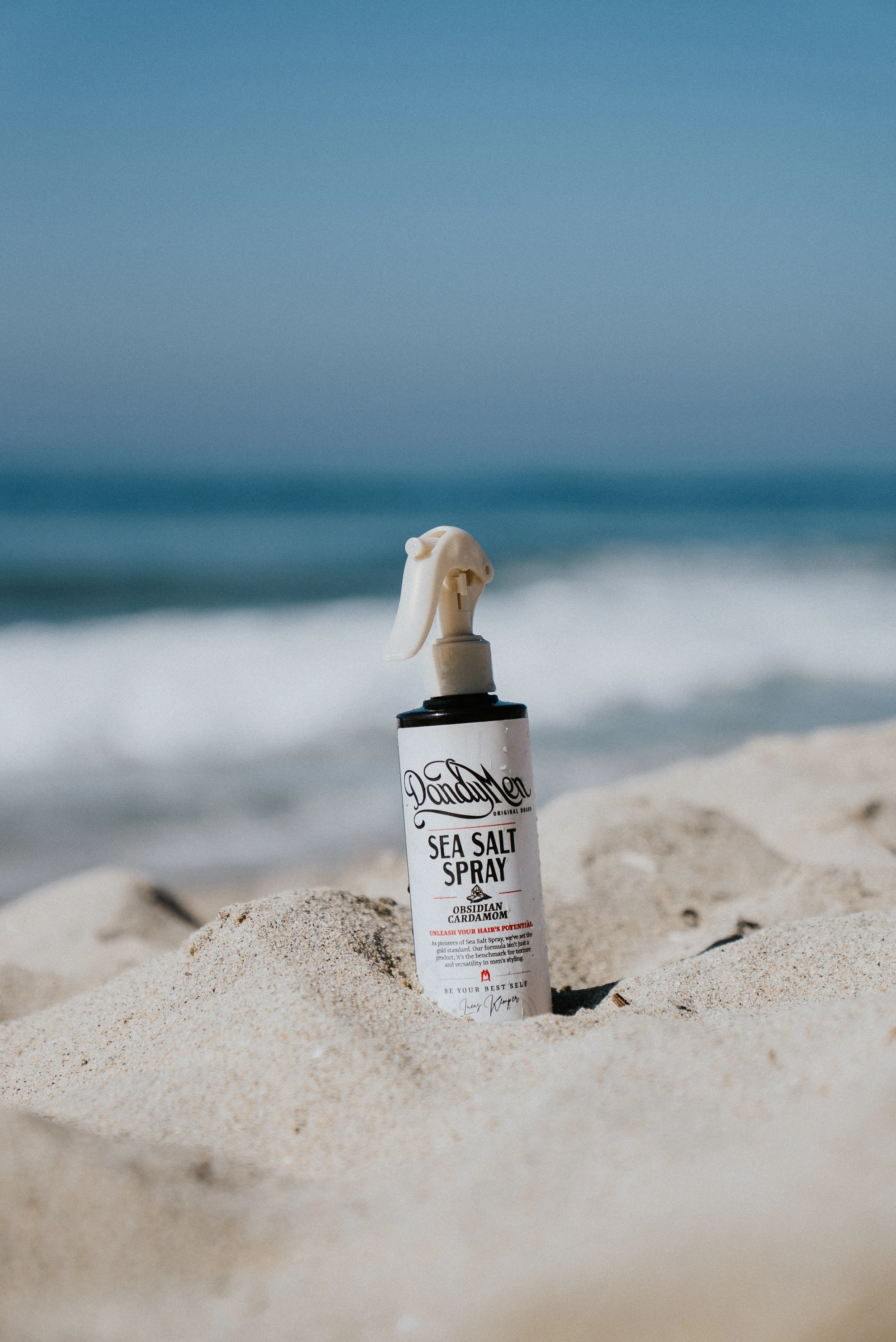 A bottle of sea salt spray with a spray nozzle, partially buried in sand on a beach, with the ocean and sky in the background.