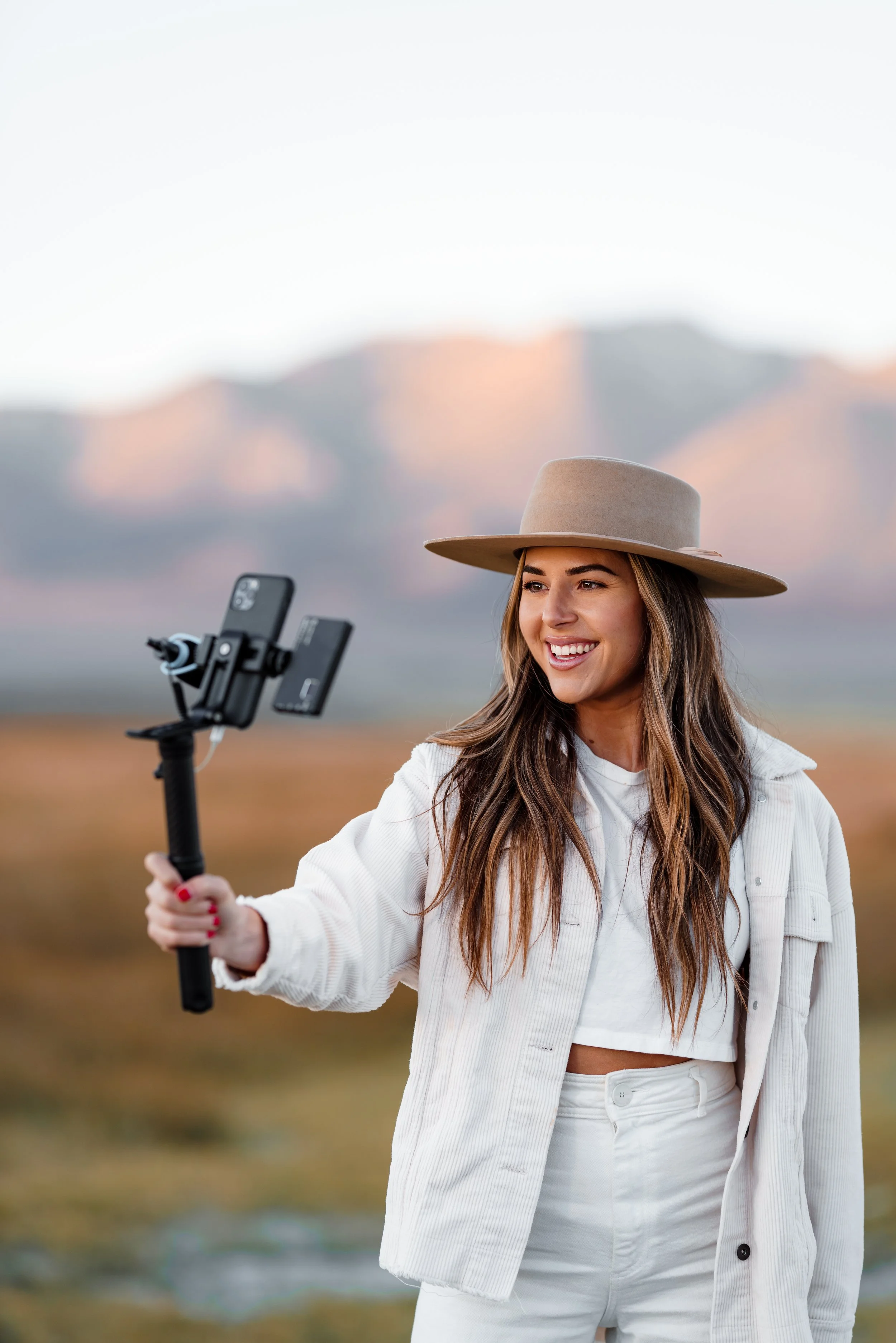 A woman outdoors wearing a beige wide-brimmed hat and white outfit, taking a selfie with a smartphone on a selfie stick, with mountains in the background.