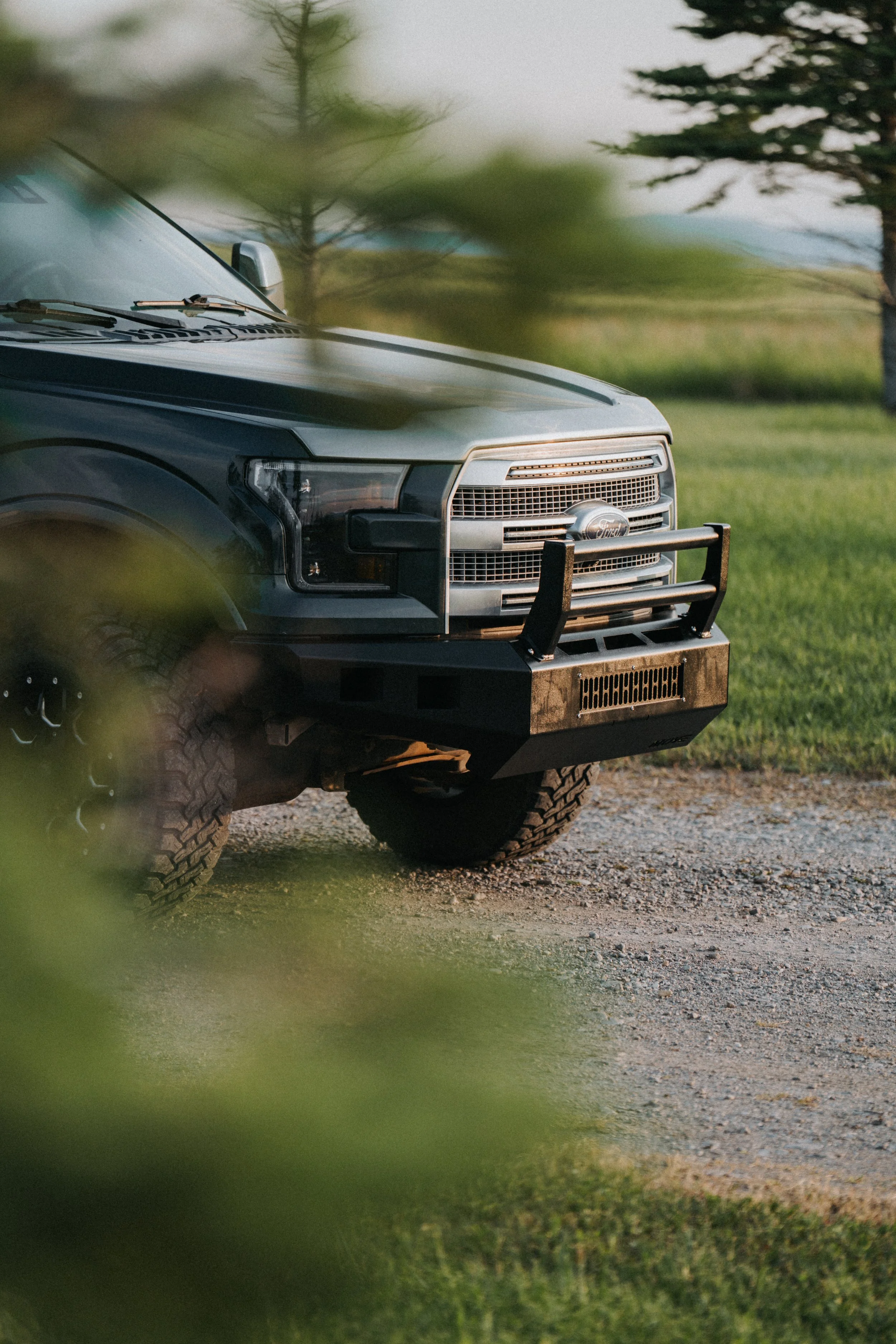 A black Ford truck with a front grille, large tires, and a push bumper, parked on a gravel surface amidst green grass and trees.
