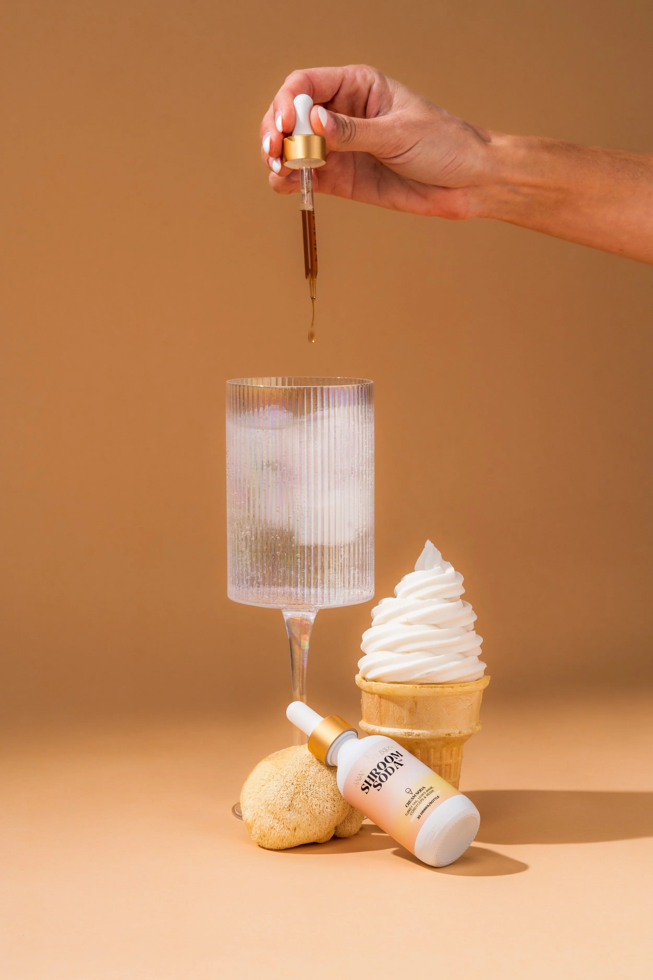 A hand holds a dropper with a brown liquid above a glass with ice. On the table, there is an ice cream cone with white soft serve, a bottle labeled 'VANISH SHROOM,' a cookie, and a tall, textured drinking glass.