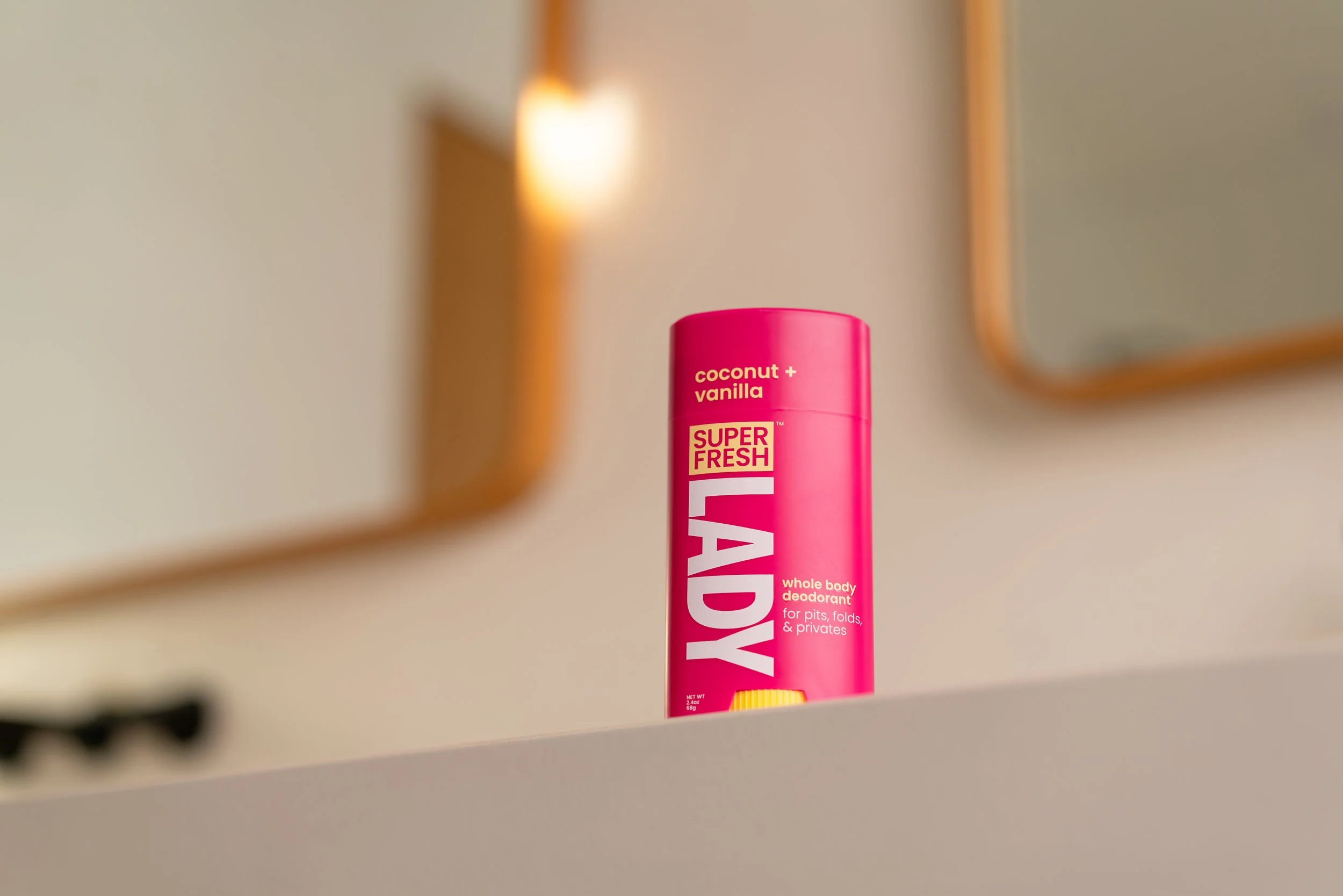 A pink spray can of Super Fresh LADY whole body deodorant with coconut and vanilla scent on a white surface, with mirrors and warm lighting in the background.