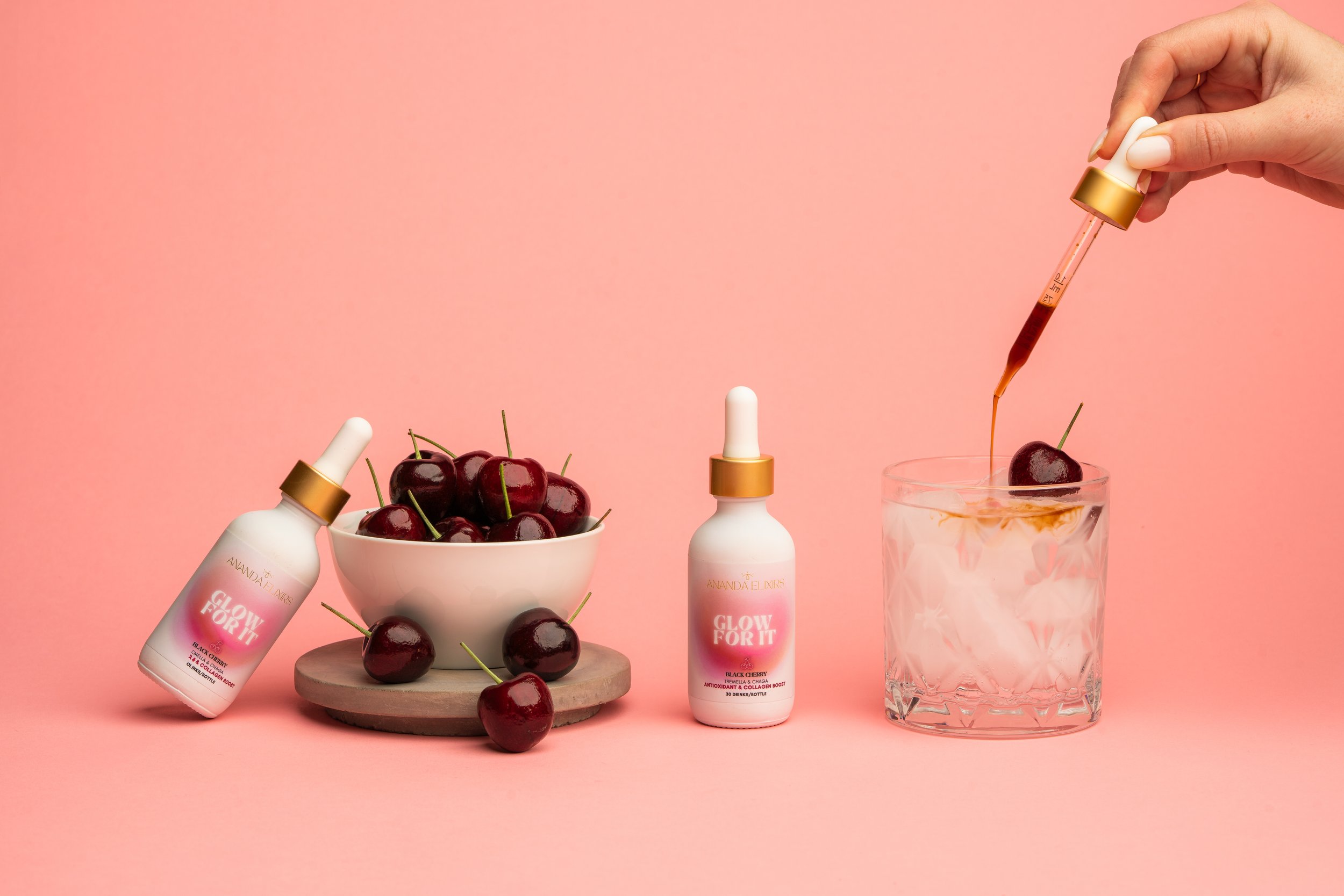 A skincare product bottle labeled 'Glow For It' placed beside a bowl of cherries and a glass of water with cherries, with a dropper held above the glass containing cherry juice