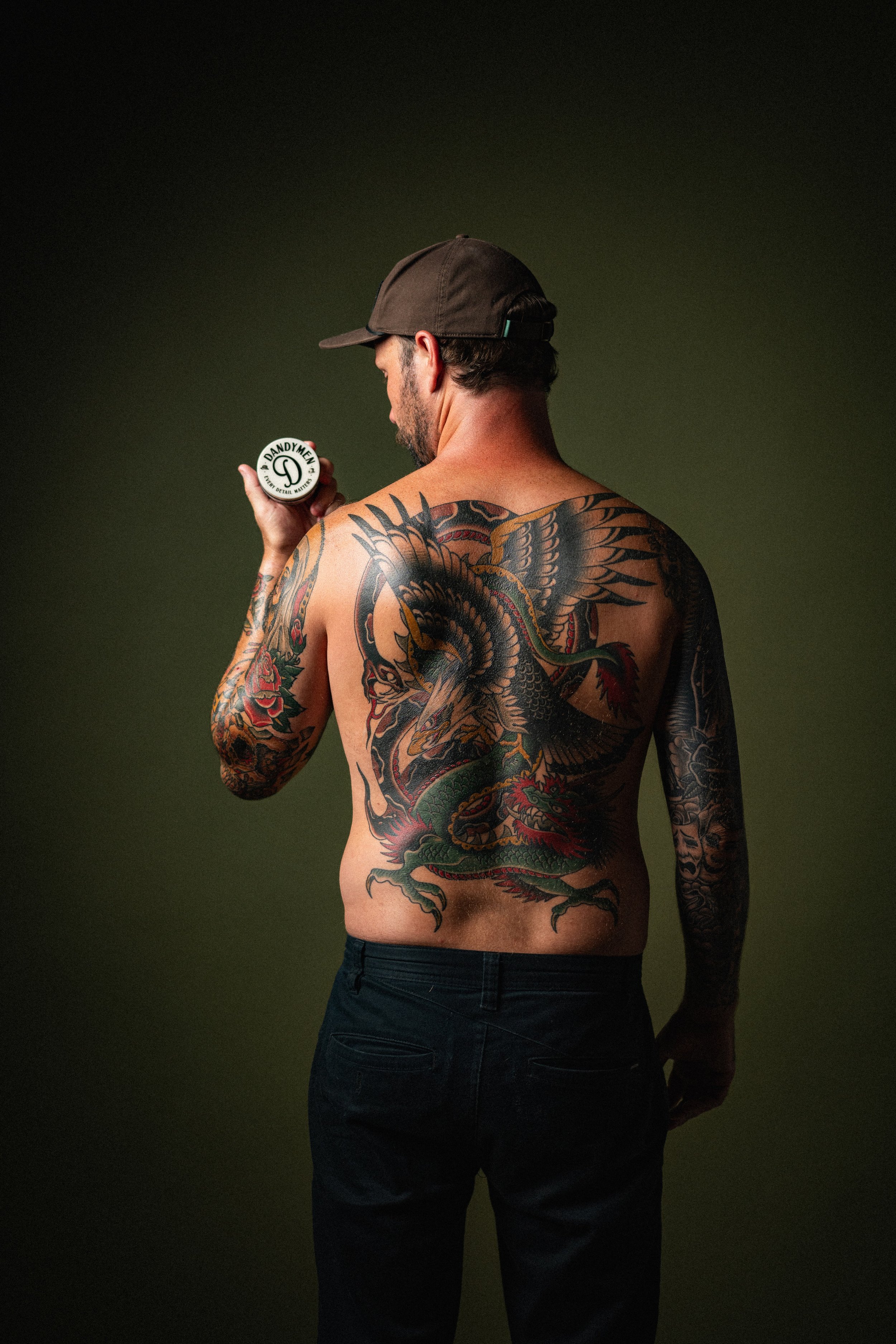Tattooed man wearing a cap and black jeans, holding a small container with the logo 'DANDYME' on it, standing against a dark green background.