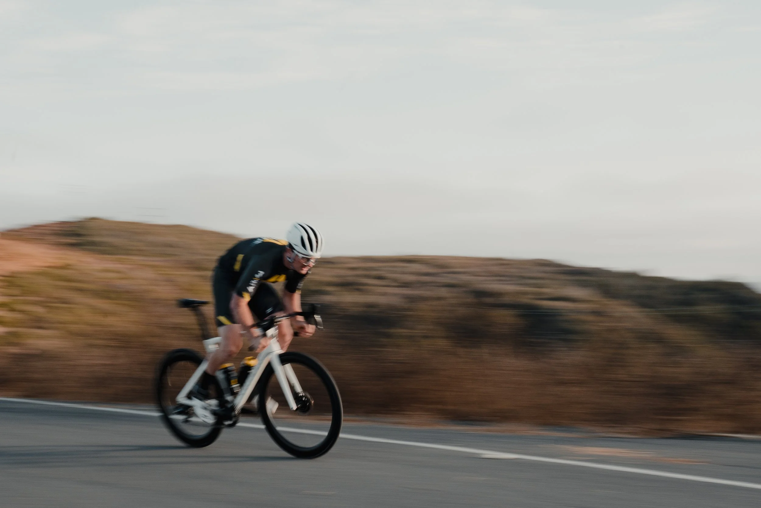 A cyclist wearing a helmet and black cycling gear riding a white mountain bike on a paved road in a hilly landscape with a cloudy sky.