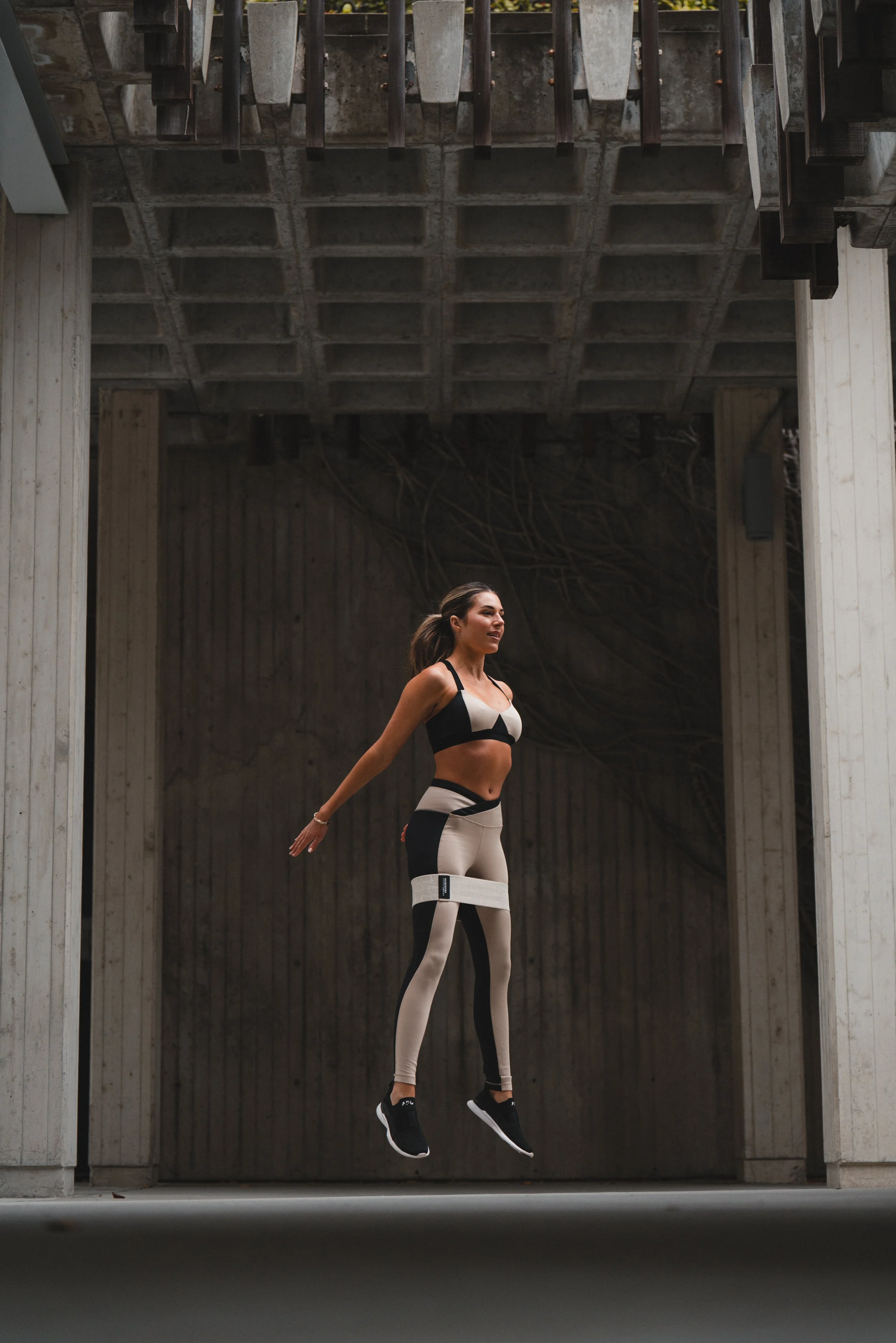 A woman in athletic wear jumps in the air outdoors, under a concrete structure with wooden elements.