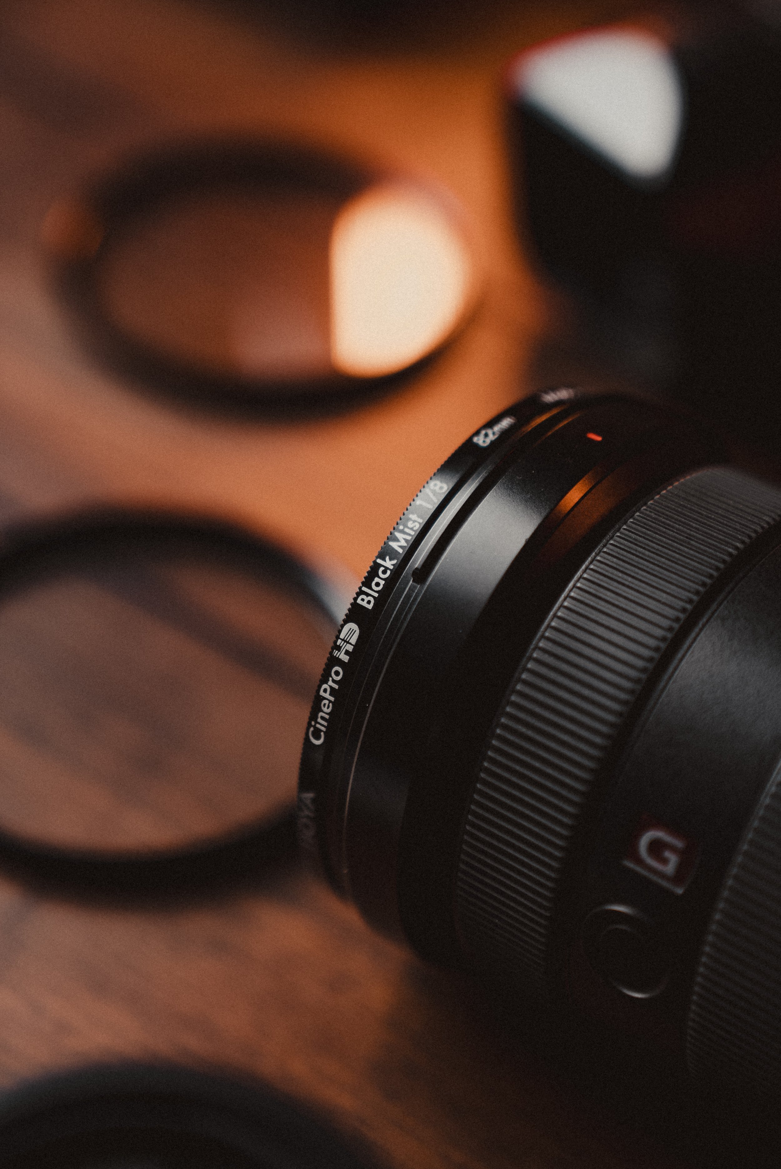 Close-up of a camera lens with focus rings, resting on a wooden surface, with additional camera lenses blurred in the background.
