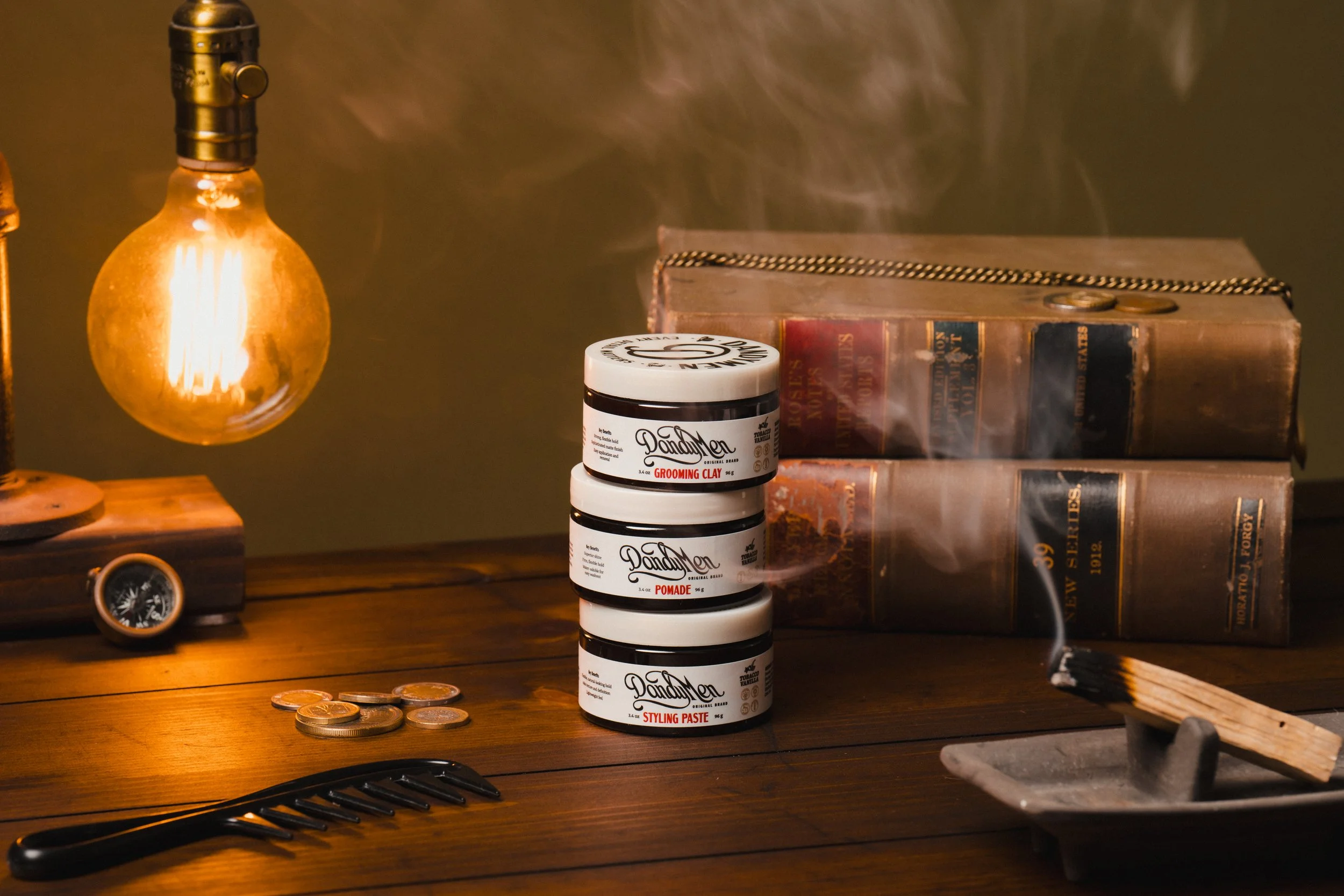 Stacked jars of beard grooming products on a wooden surface, with stacked old books in the background and a lit matchstick emitting smoke nearby.