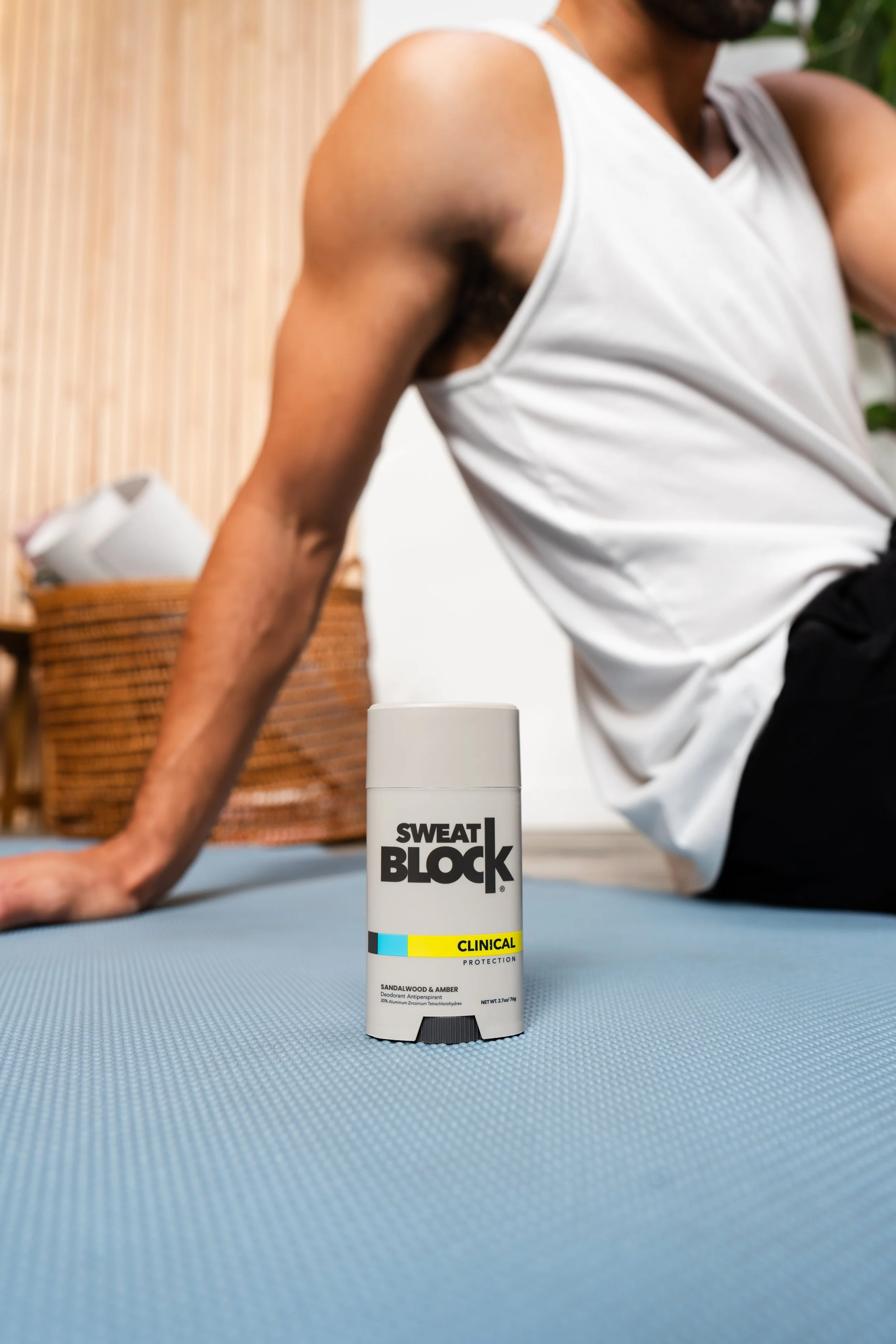 A container of Sweat Block clinical deodorant on a blue exercise mat, with a man in a white sleeveless shirt sitting on the floor behind it.